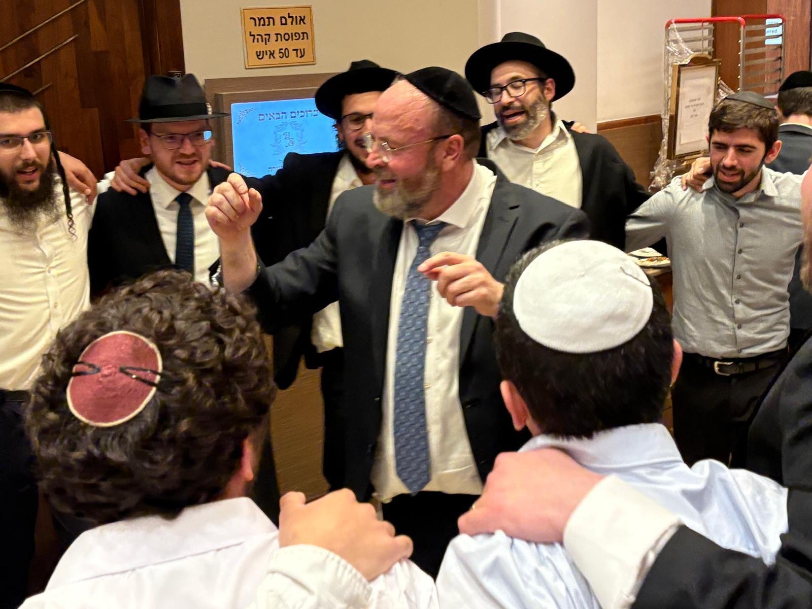 A group of Jewish men wearing kippahs and hats are gathered in a circle, smiling and celebrating together at a party or celebration.