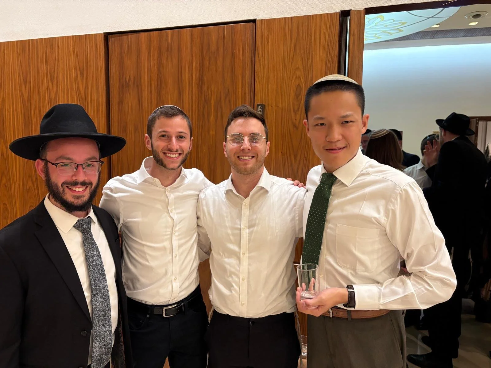 Four men standing together at an indoor social event, dressed in formal attire, smiling at the camera. One man is wearing a black hat and glasses, holding a glass; others are in white shirts, with one wearing a tie and another wearing a kippah.