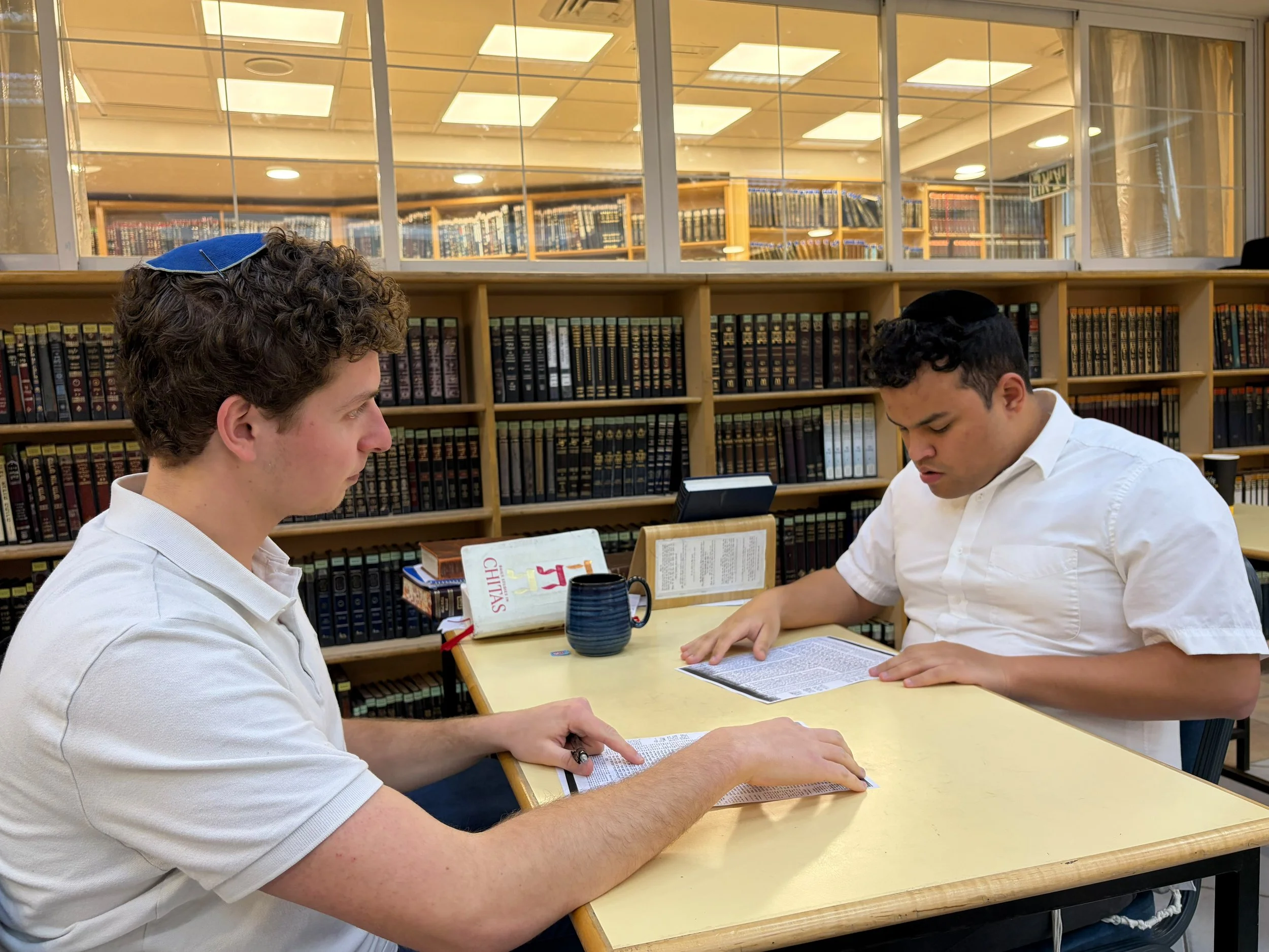 Two young men wearing white shirts are sitting at a yellow table in a library. One is reading a book, and the other is looking at a smartphone. Behind them are shelves filled with books, and a window with a view of another room with more bookshelves.