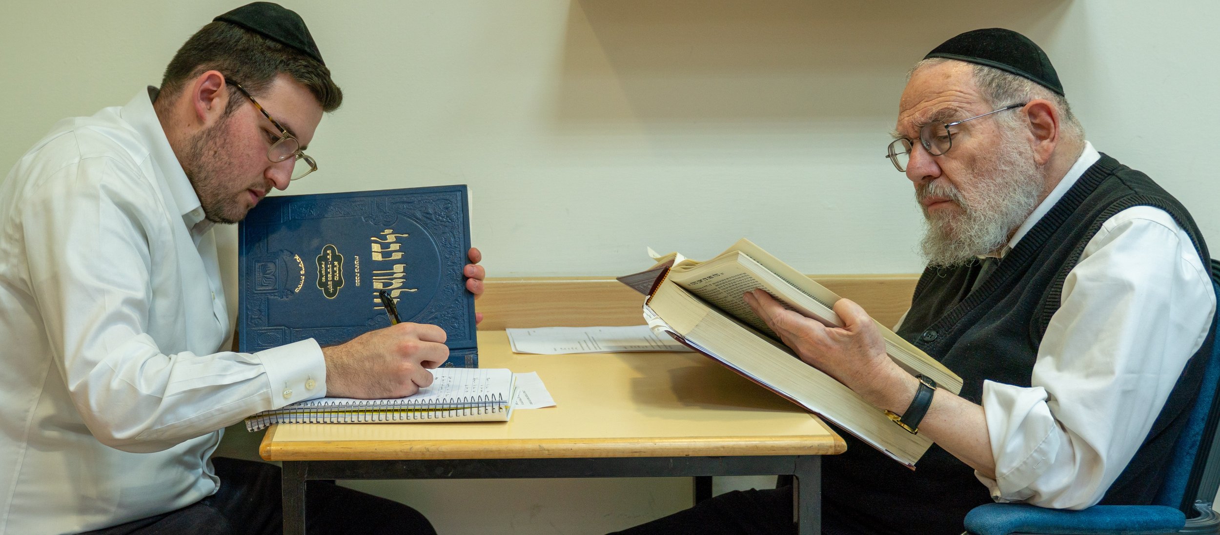 Two men wearing Jewish prayer hats are seated at a table, reading religious texts and taking notes.