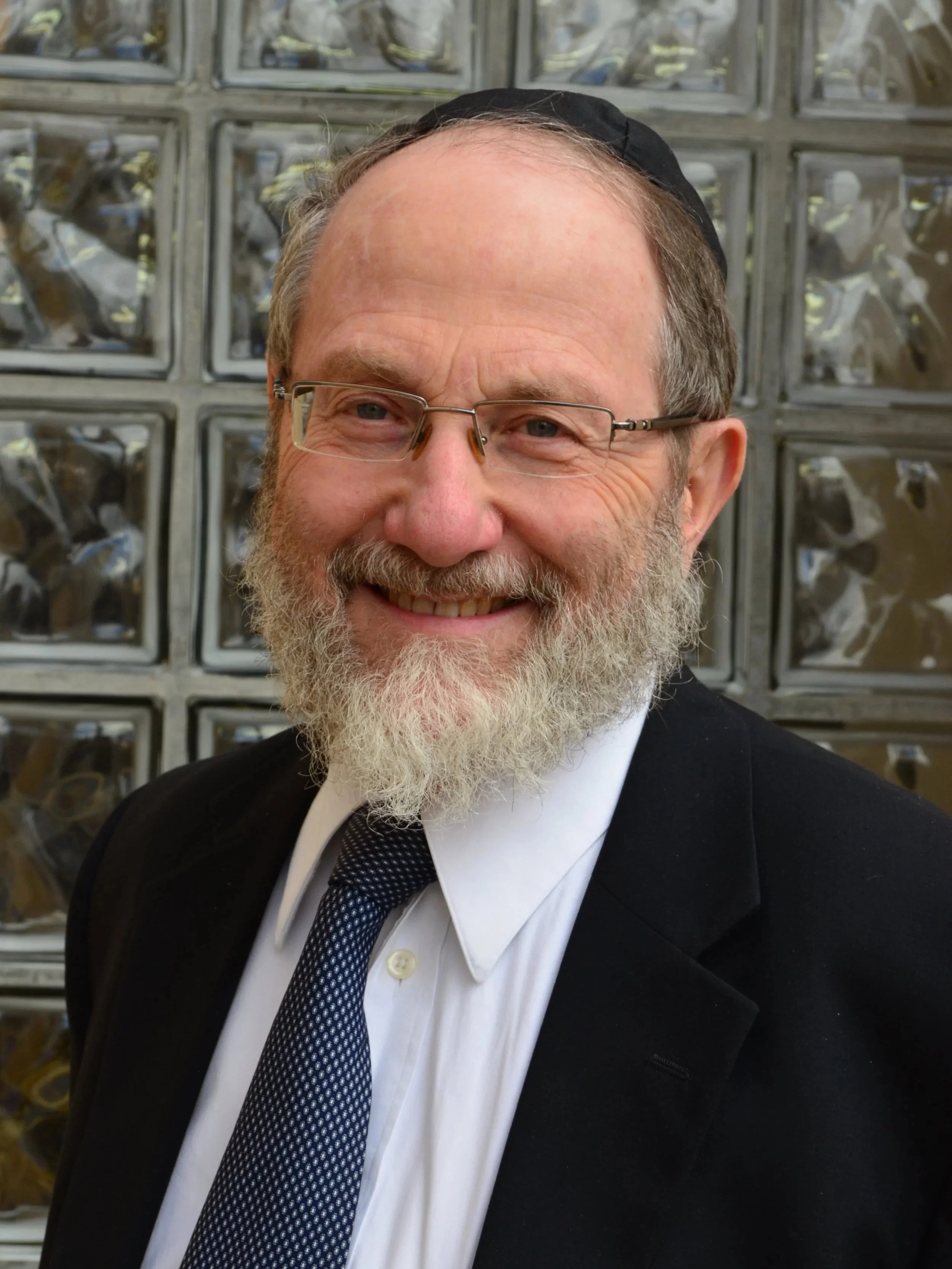 A smiling middle-aged man with glasses, a beard, and a kippah in a black suit, white shirt, and patterned tie, standing in front of a wall of glass blocks.
