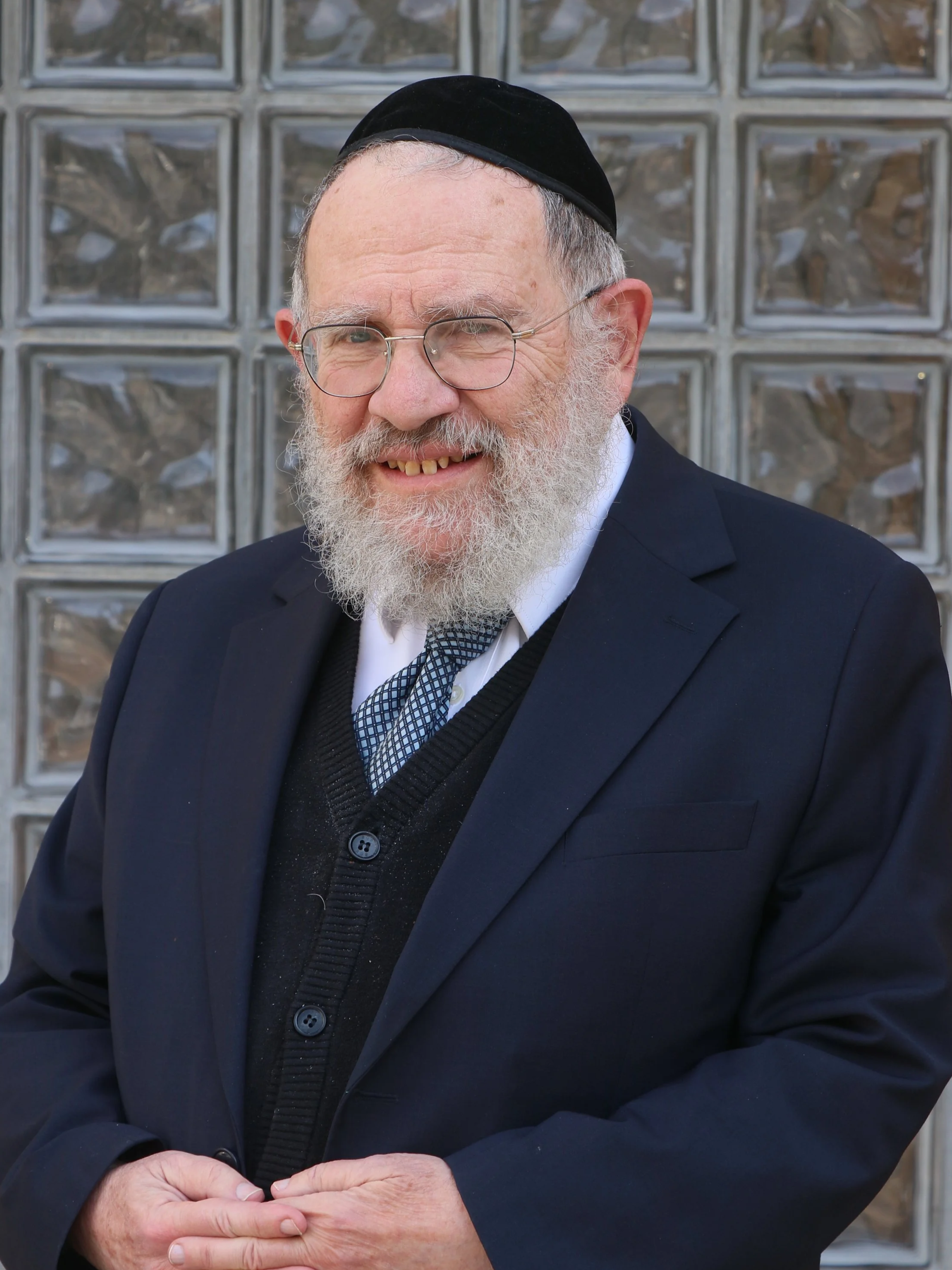 A senior man with glasses, a white beard, and a black yarmulke, dressed in a black suit and tie, smiling and clasping his hands in front of him, standing in front of a glass brick wall.