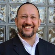 A smiling man with a beard and glasses, wearing a dark blazer and a white shirt, standing outside in front of a glass block wall.