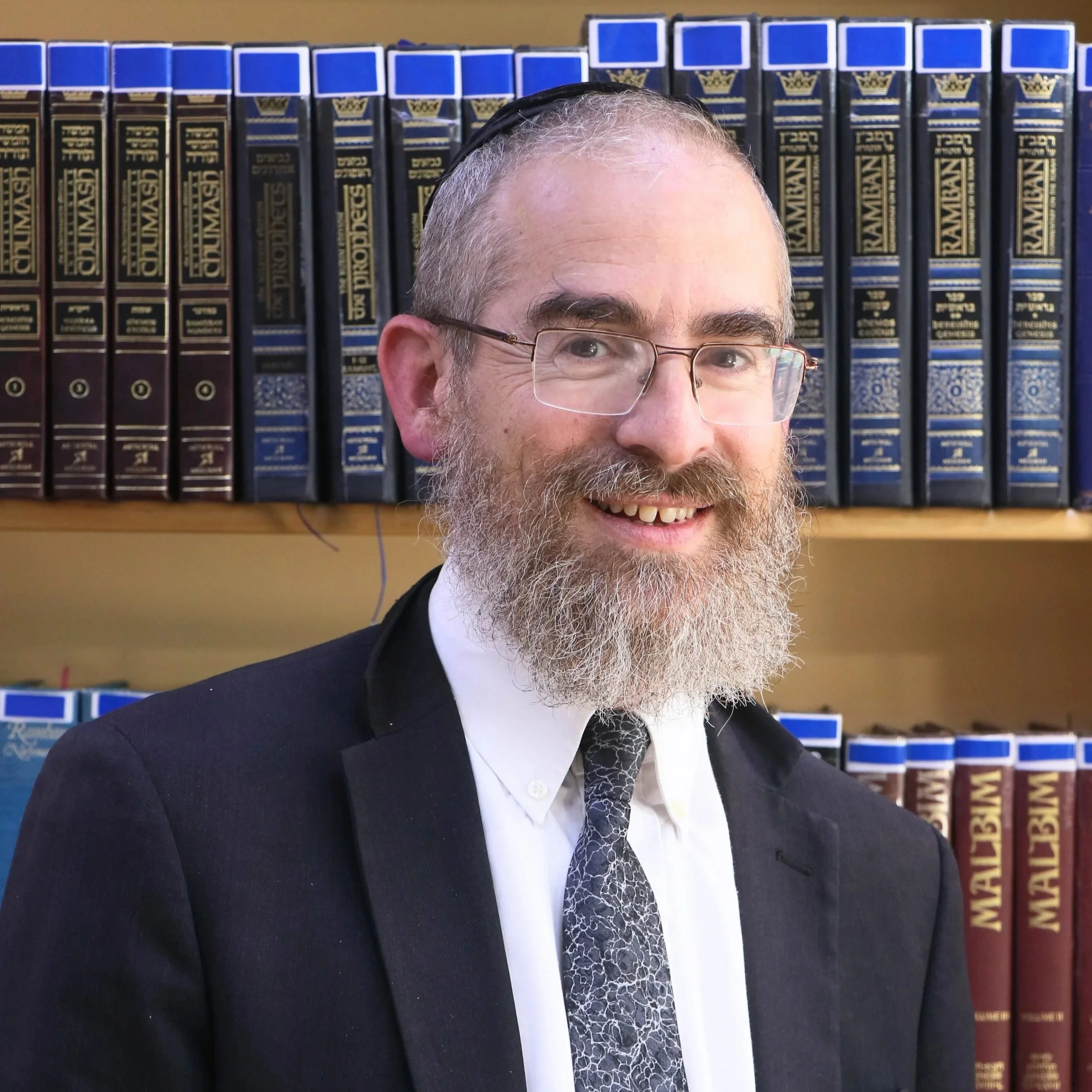 Close-up of a smiling man with glasses, beard, and gray hair wearing a black suit and patterned tie, standing in front of a bookshelf filled with law books.