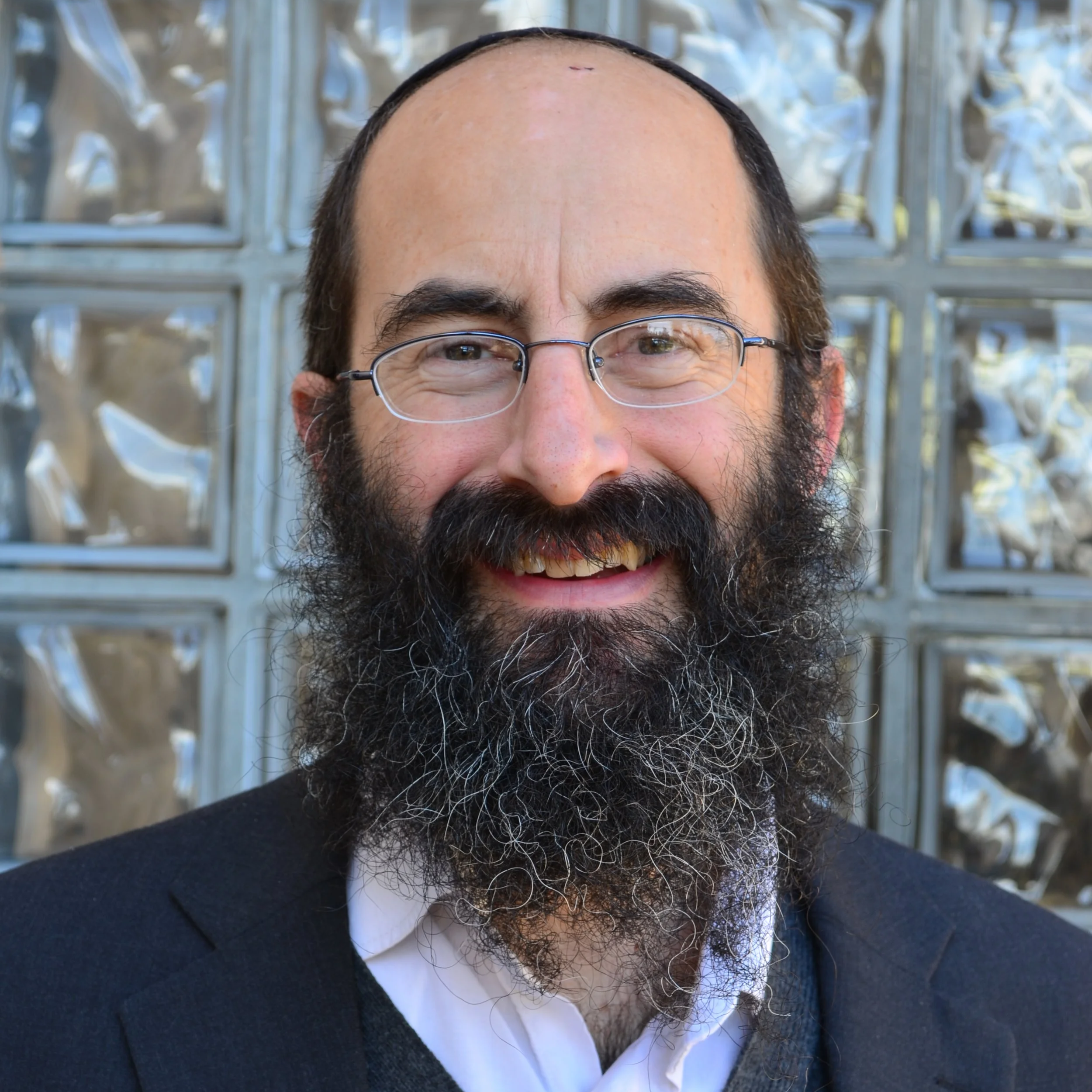 Close-up of a smiling man with glasses, dark beard, and hair, wearing a dark sweater and white shirt, standing outdoors in front of a metal lattice structure.