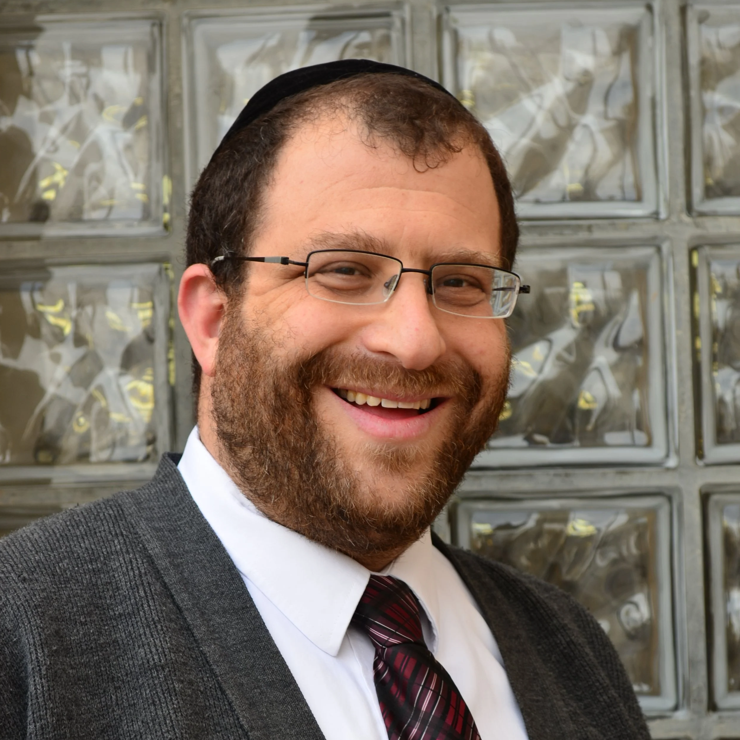 Close-up of a smiling man wearing glasses, a suit, a white shirt, and a striped tie, with a glass block wall in the background.