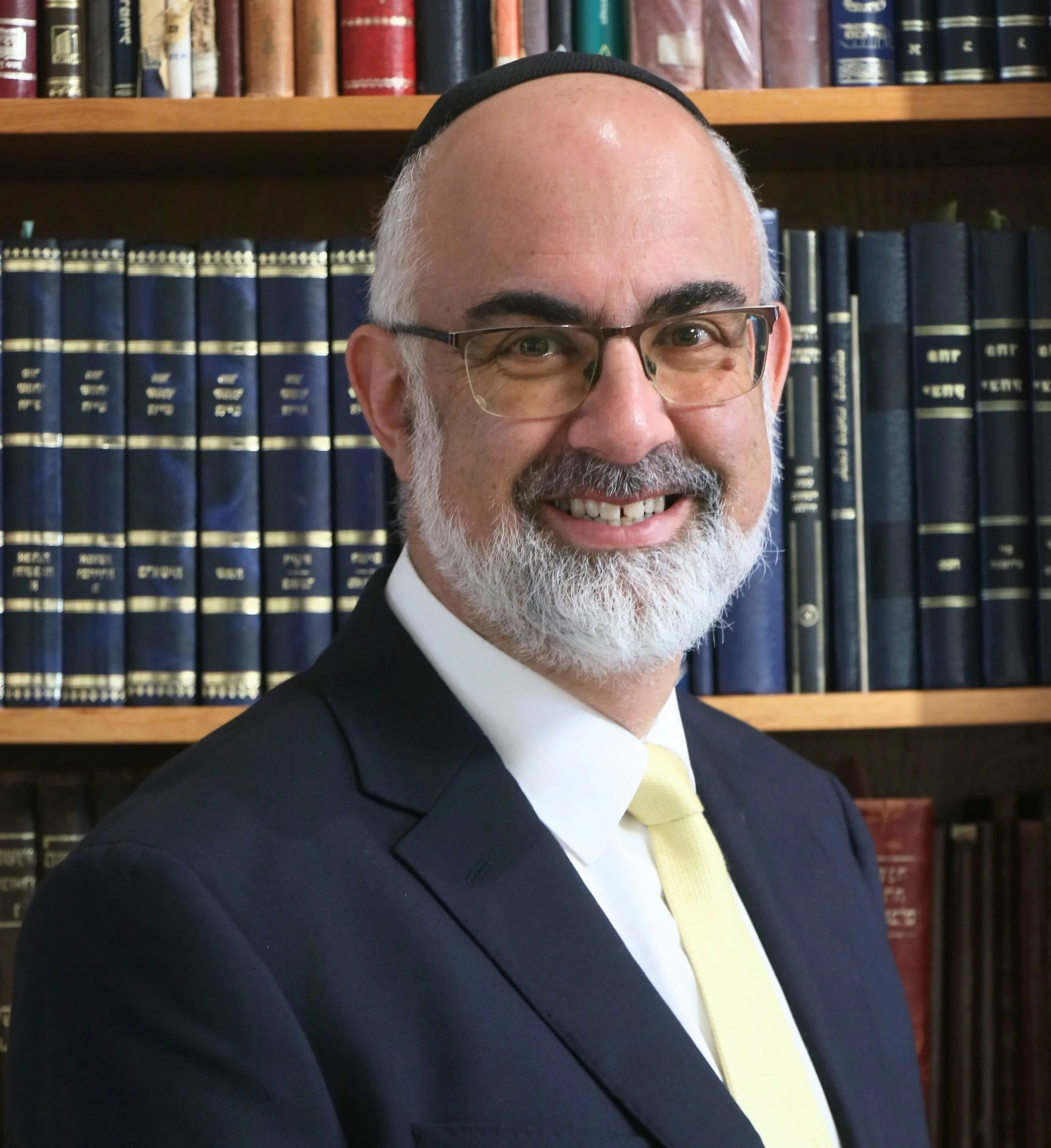 Close-up of smiling man with glasses, wearing a yarmulke, in front of a bookshelf filled with Jewish religious books.