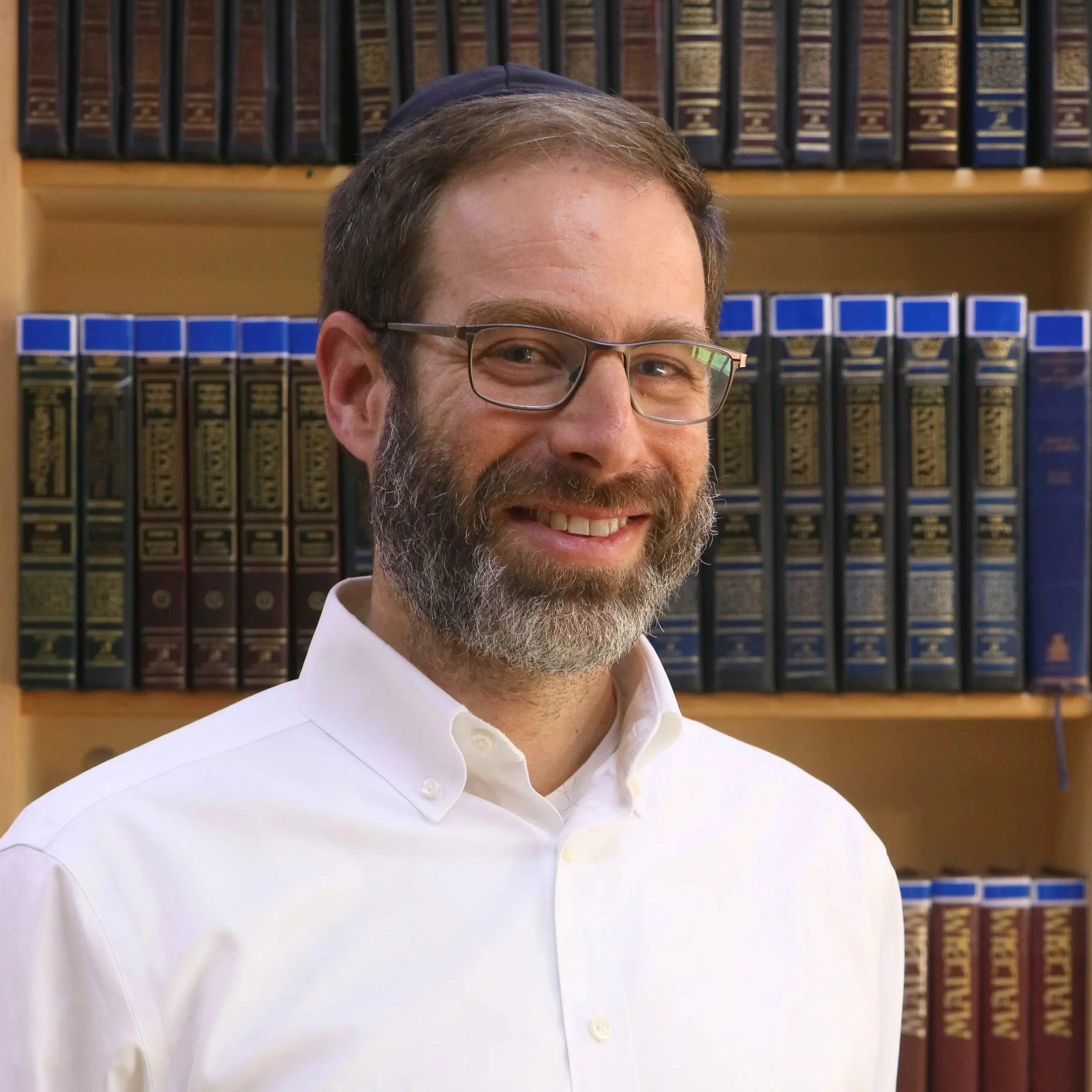 A smiling man with gray hair, beard, and glasses, wearing a white shirt, standing in front of a bookshelf filled with legal or reference books.