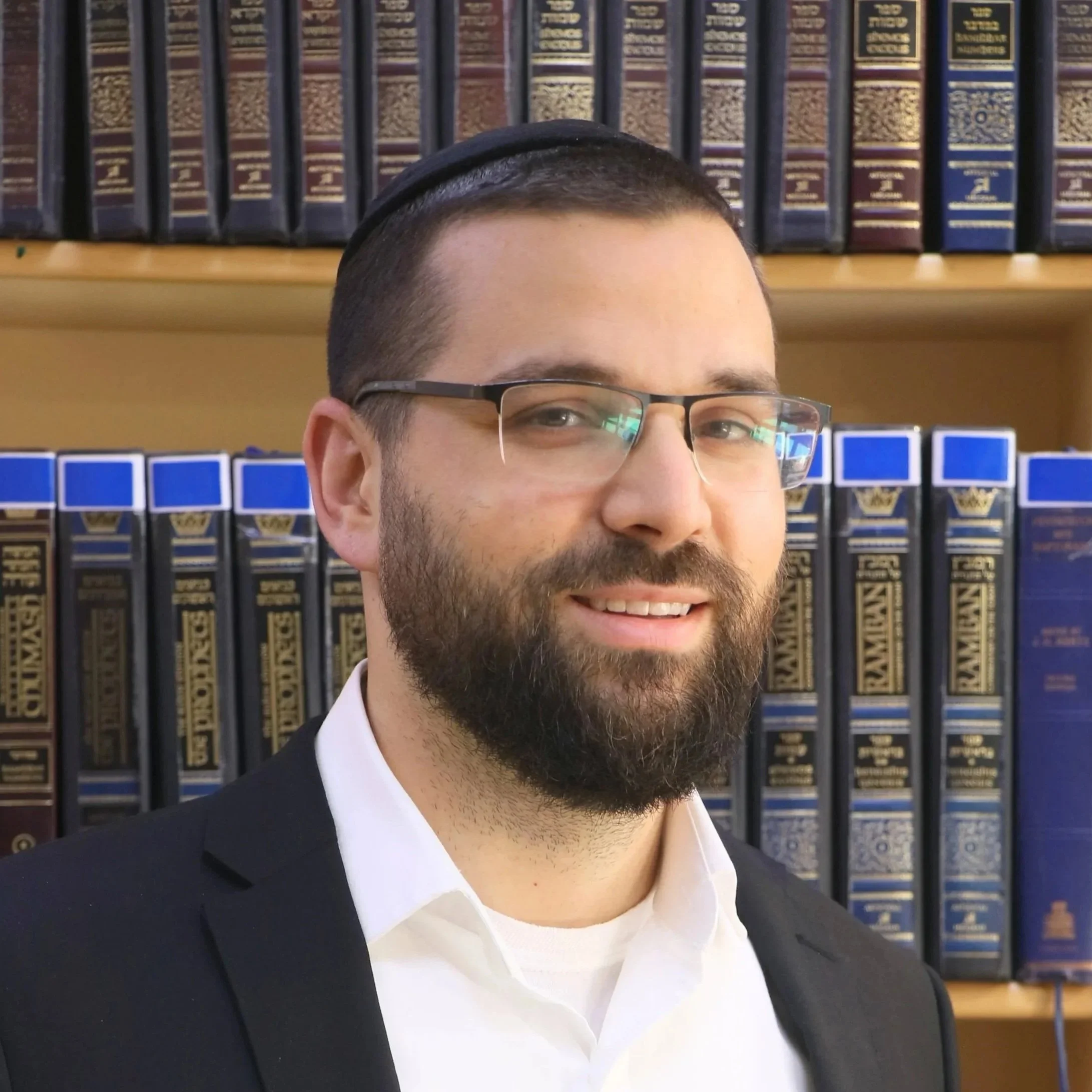 Close-up of a man in a suit and white shirt, wearing glasses and a yarmulke, smiling, with a bookshelf filled with colored and labeled books in the background.