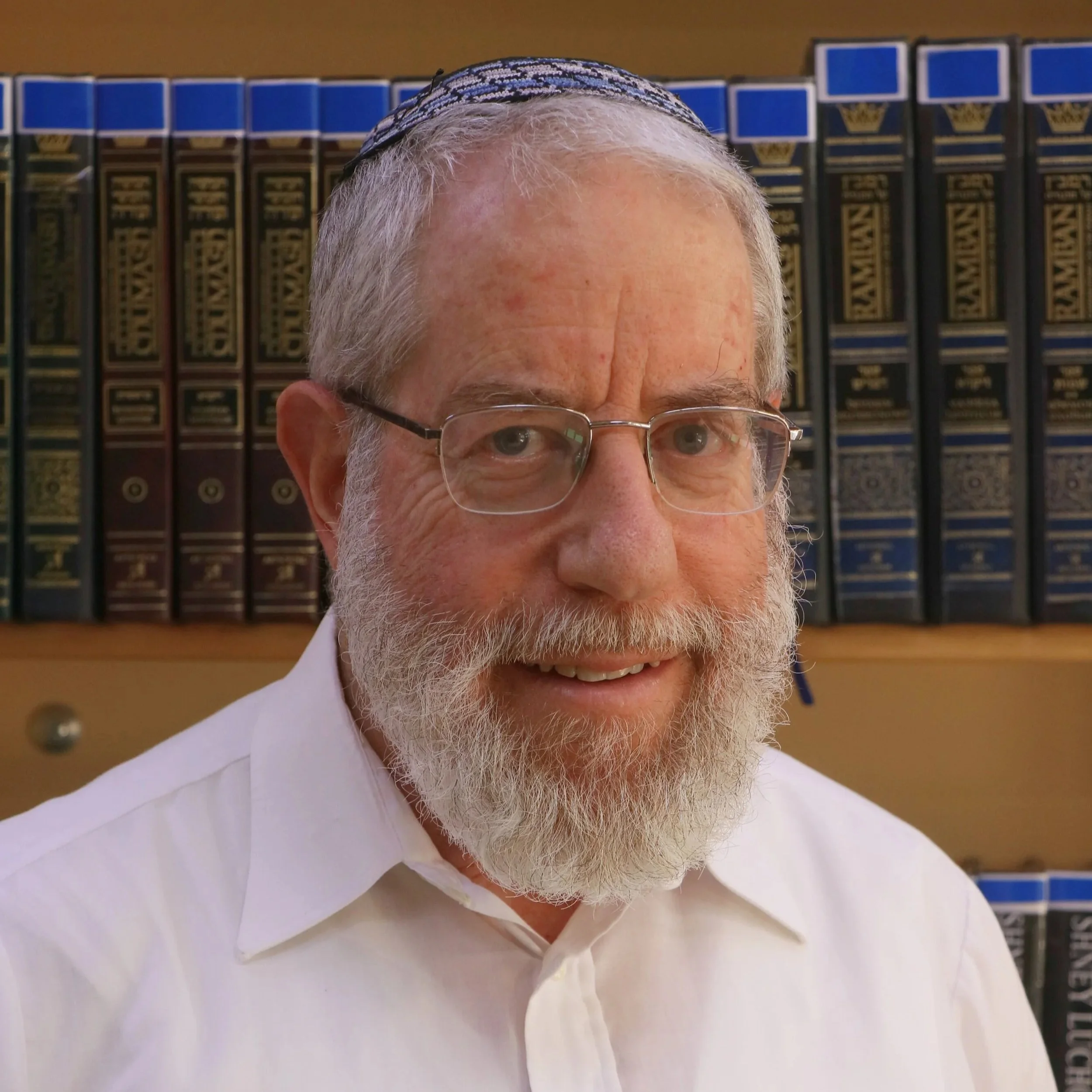 Close-up of an older man with gray hair, beard, glasses, wearing a white shirt, smiling, with bookshelves in the background.