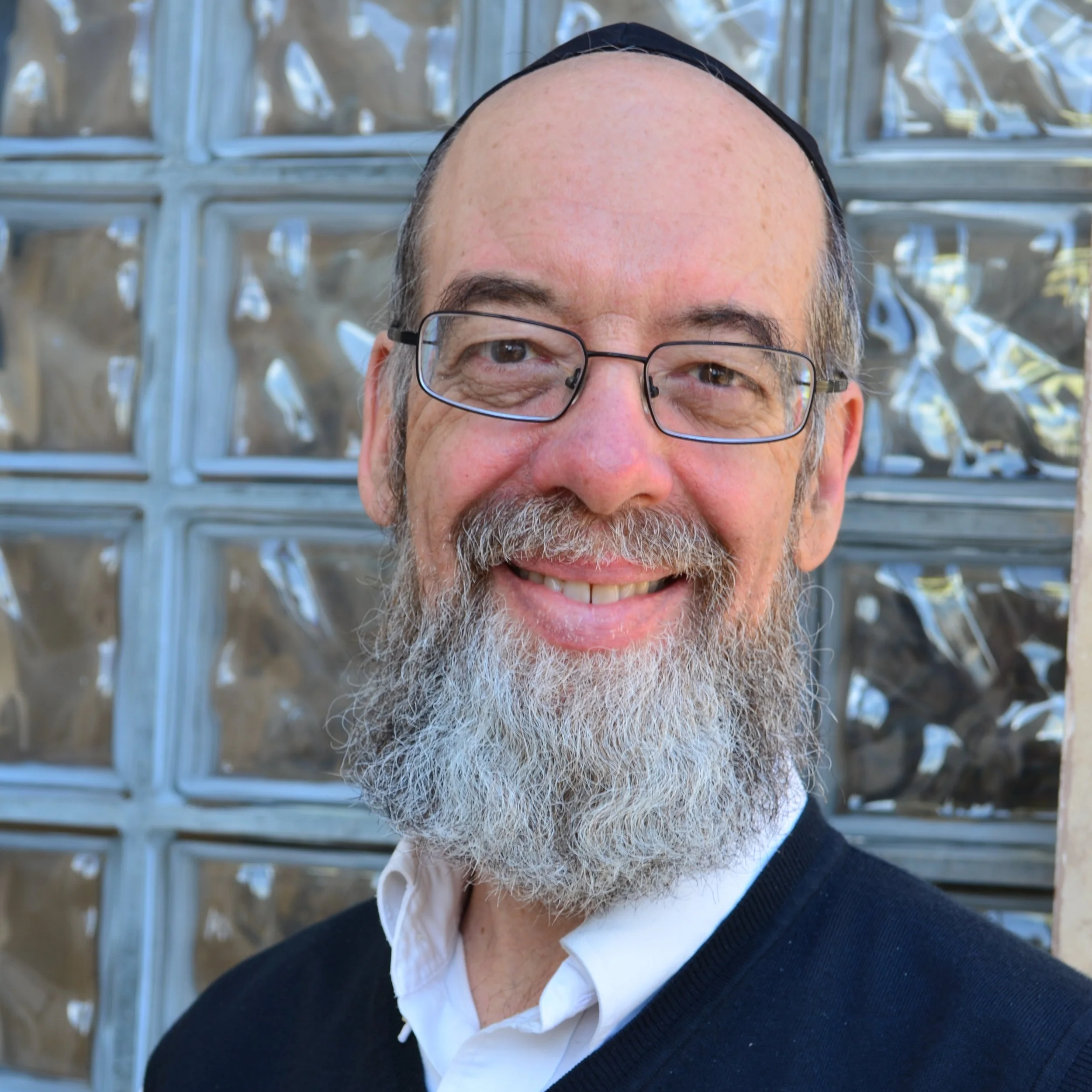 Close-up of a smiling man with glasses, partially balding, with a beard, wearing a black sweater and white collared shirt, standing outdoors in front of a metal fence.