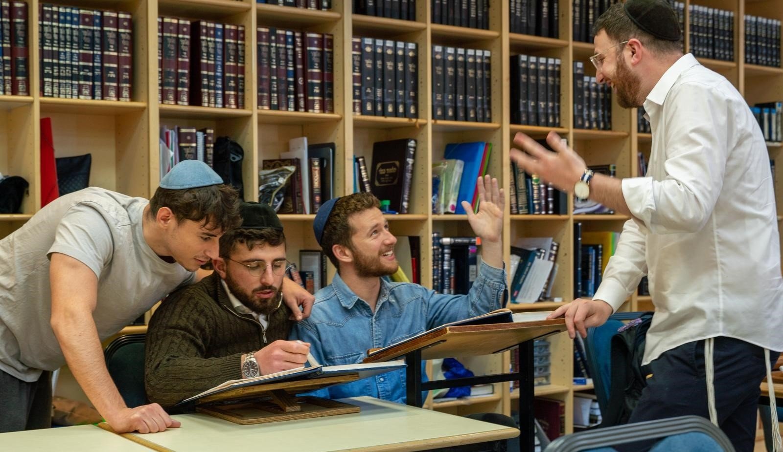 Four men, some wearing yarmulkes, engaged in lively discussion or study in a room with a wooden bookshelf filled with books behind them. One man is standing, gesturing with his hands while the others sit at a table, some looking at notebooks or books.