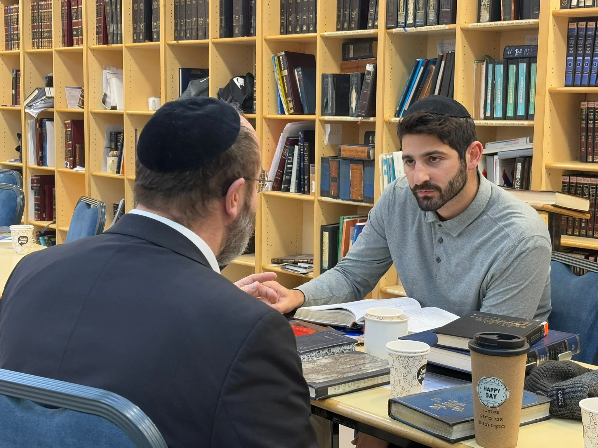 Two men, one with a beard and glasses wearing a kippah and a suit, and the other with dark hair and a beard wearing a jacket, are sitting across from each other at a table, engaged in conversation in a library or study room with wooden bookshelves filled with books behind them. The table has open books, papers, and disposable coffee cups.