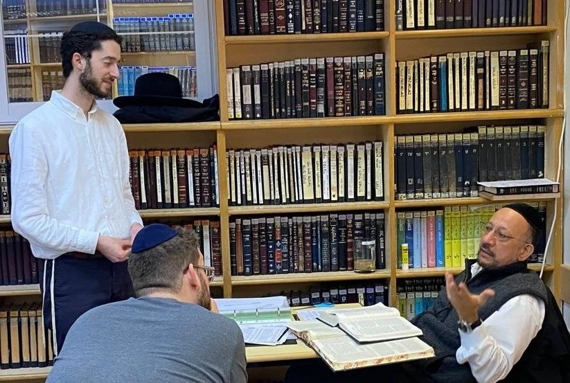 Three men in a library or study room, engaged in conversation, with bookshelves filled with books in the background. One man with glasses and a kippah is speaking, another man is listening attentively, and a third man is sitting at the table with open books and papers.