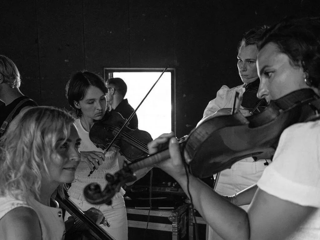 Group of women playing violins in a rehearsal room, with one woman smiling and the others focused on their instruments.