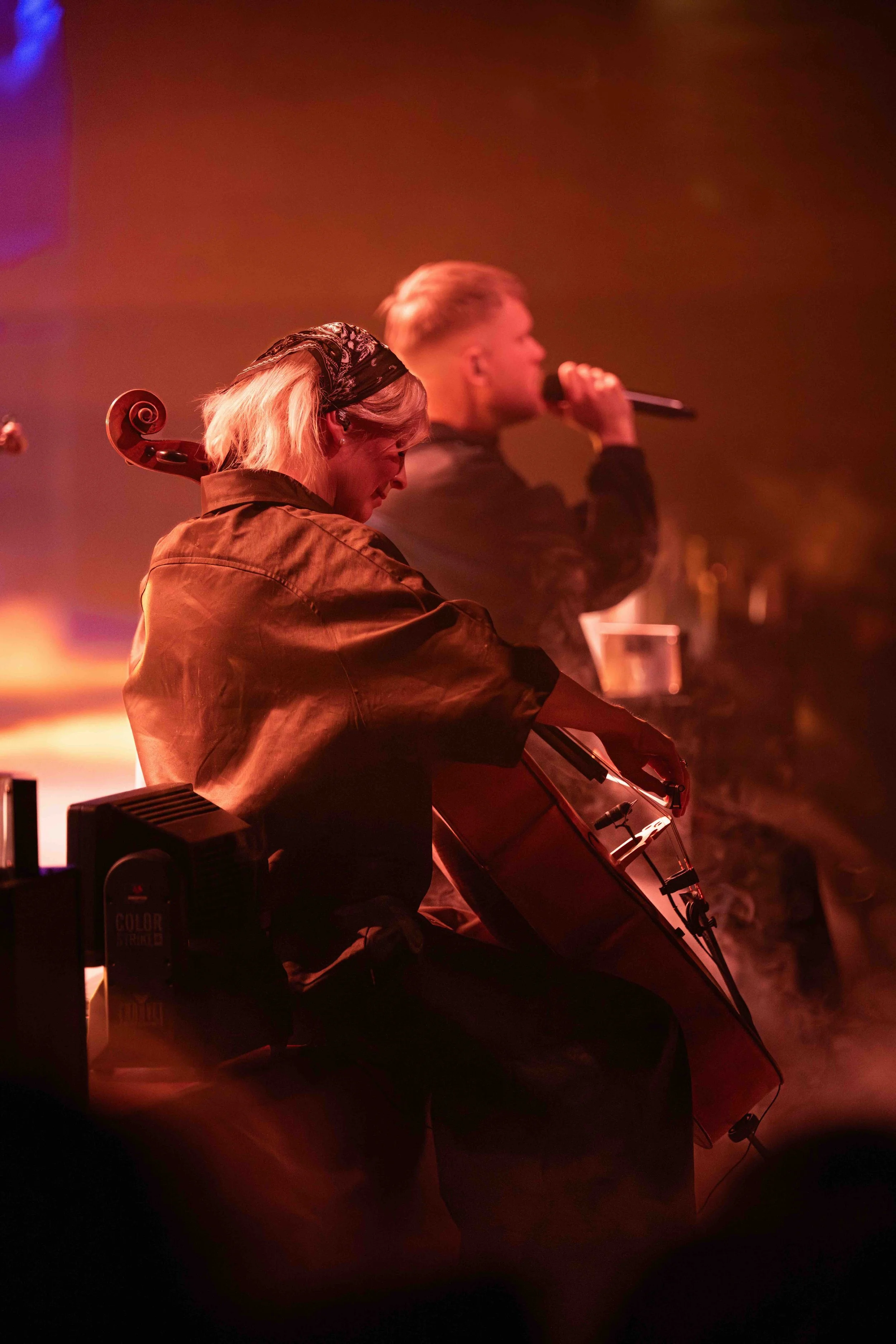 A woman with blonde hair and a headscarf sits playing a cello, while a man with short hair sings into a microphone in the background. The scene is dimly lit with warm, reddish-orange lighting, creating an intimate atmosphere.