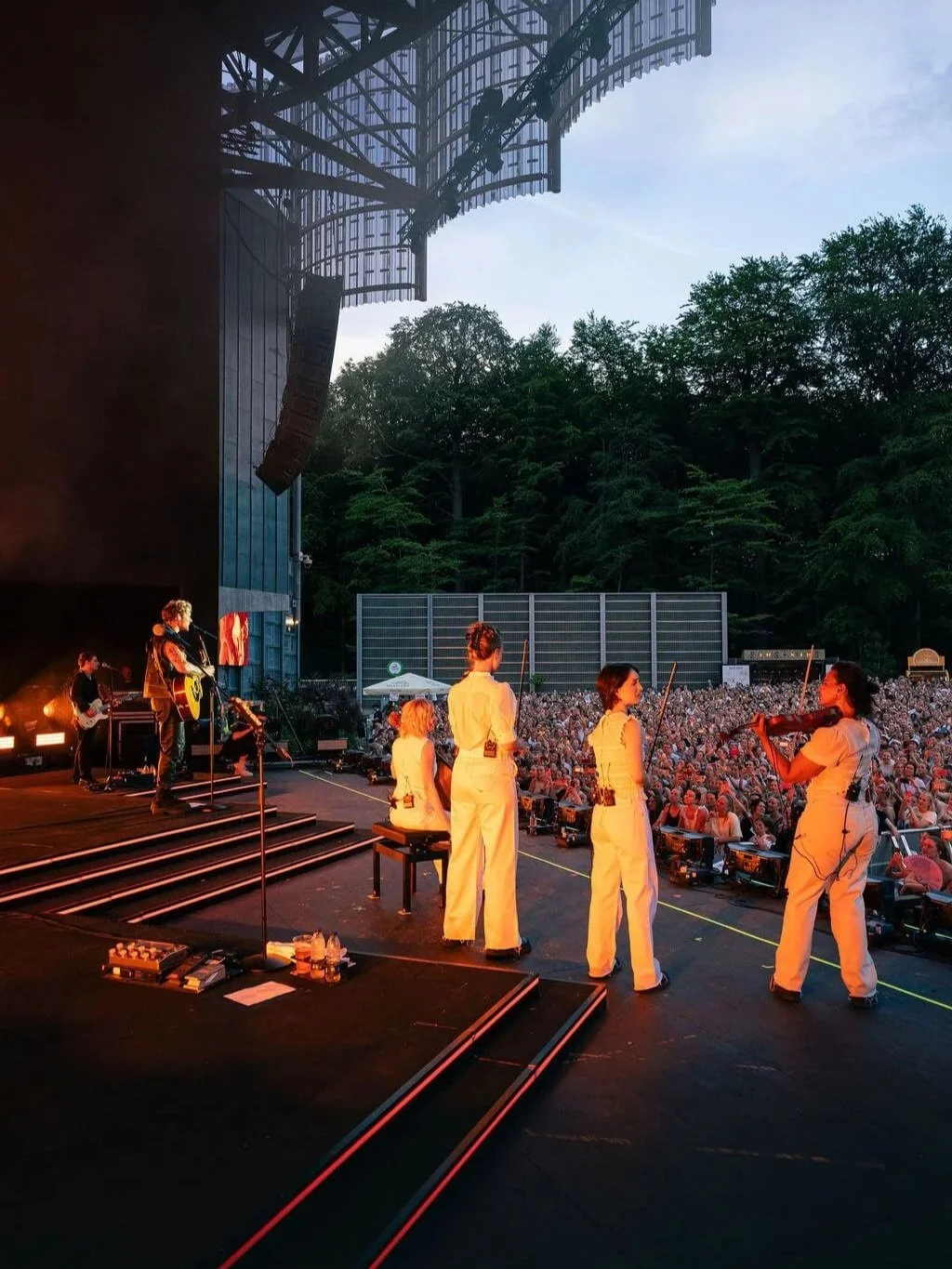 A live outdoor music performance on a stage with a band and singer, during sunset, with concertgoers in the audience and a wooded background.