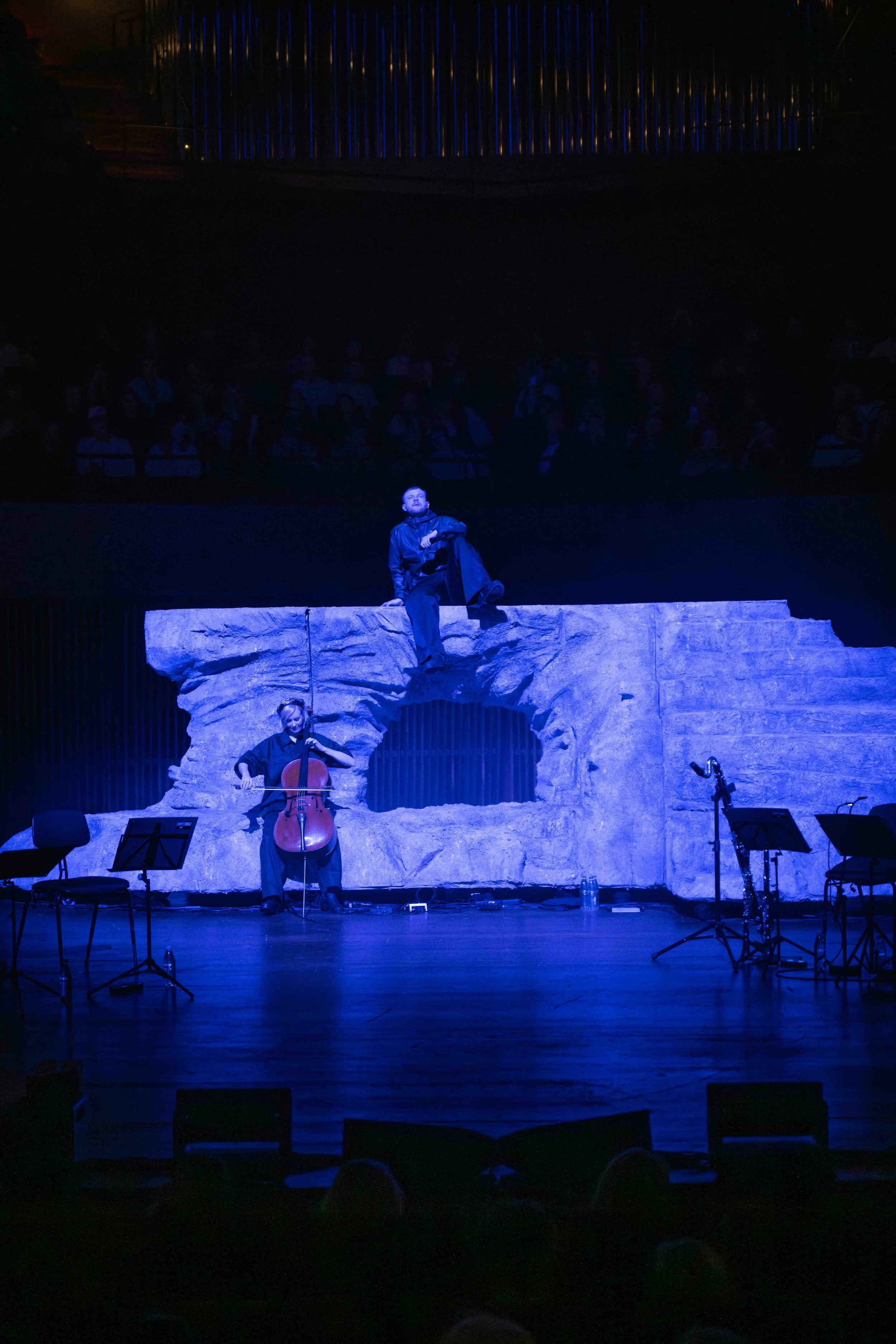 A stage with a rock formation backdrop illuminated with blue lighting, featuring a musician sitting on a ledge at the top, another sitting below with a cello, and empty chairs and music stands in the foreground.