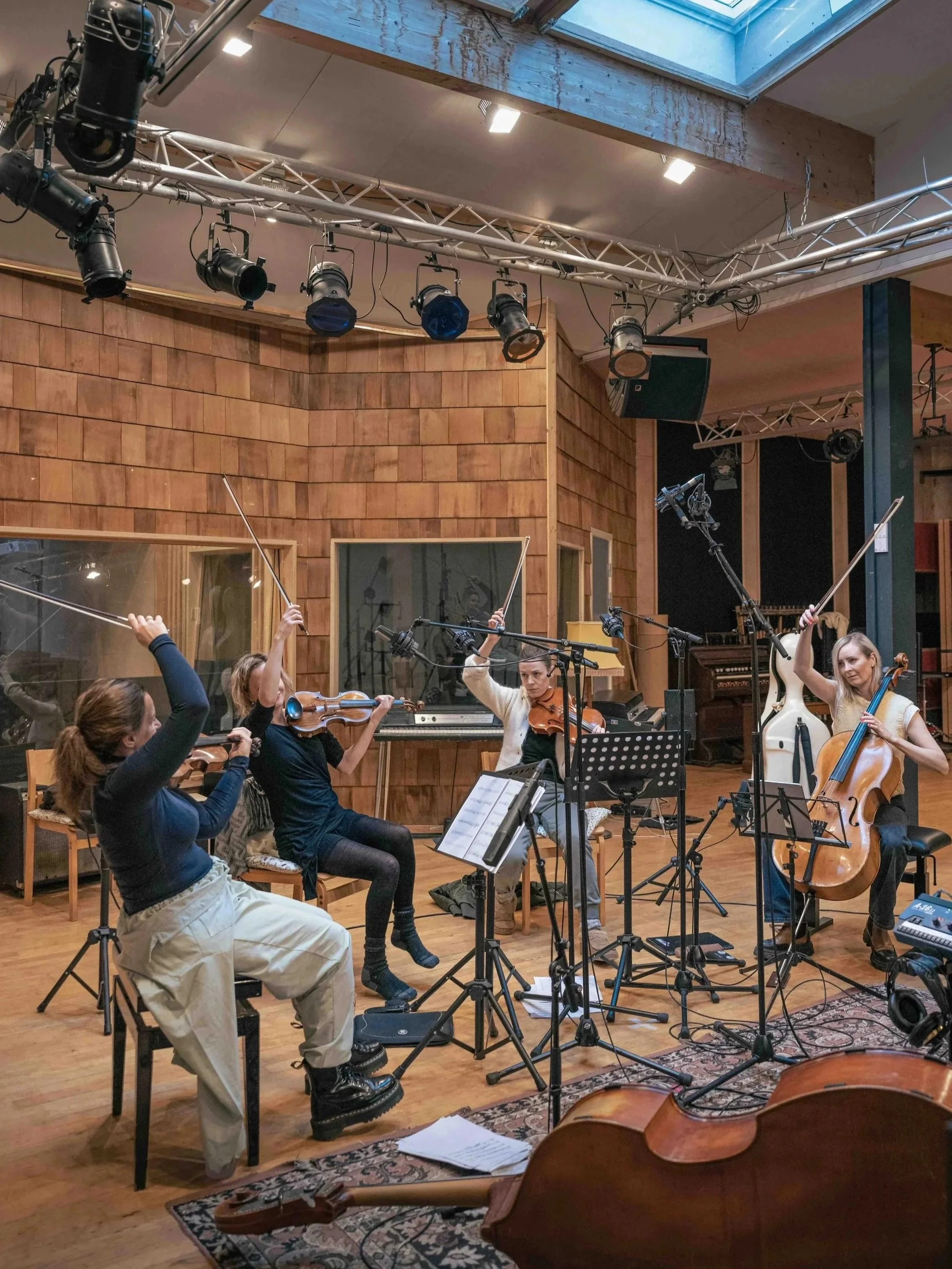 A group of musicians in a recording studio, with several of them playing string instruments such as violins and a cello. They are surrounded by music stands and microphones, and appear to be rehearsing or recording. The studio has wood-paneled walls, large windows, and stage lighting equipment overhead.