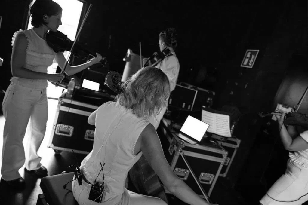Four women musicians practicing with violins and a guitar in a dimly lit room.