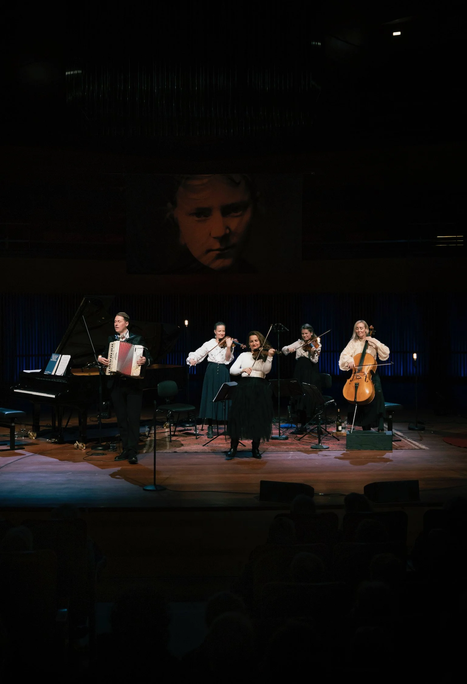 A musical performance on a stage with five classical musicians, including a pianist, accordion player, and violin and cello players, under a large portrait of a man's face.