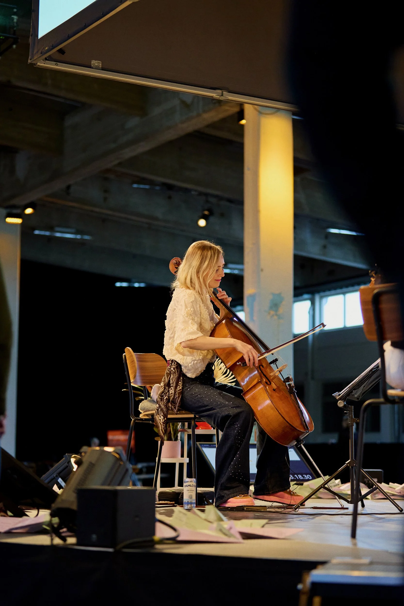 A woman with blonde hair wearing a cream-colored blouse and black pants is playing a cello on a stage, with music stands and electronic equipment around her, under a large indoor structure with industrial beams and lighting.