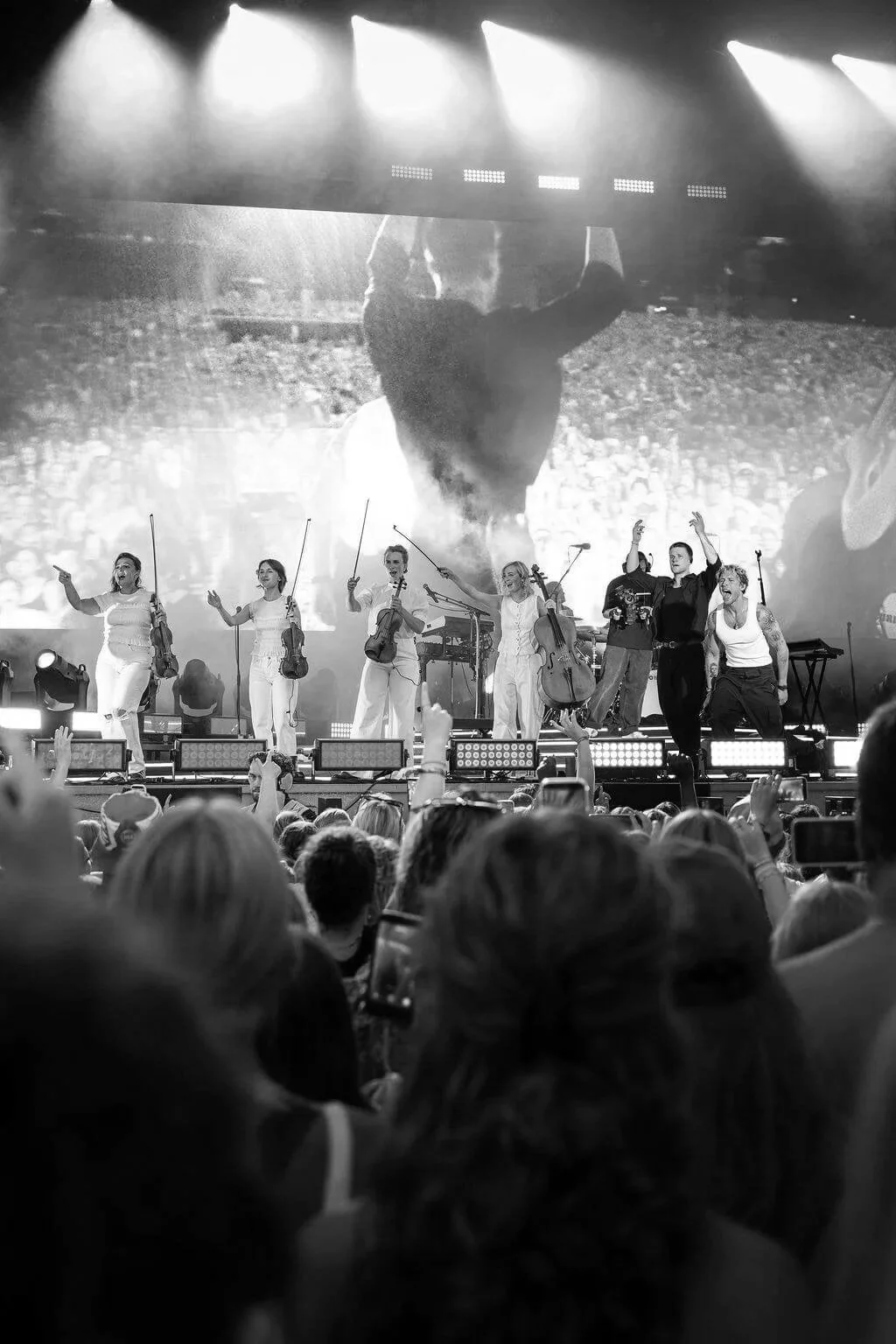 A black and white photo of a concert stage with musicians and performers, with a large screen in the background showing a silhouette of a person with their hands raised. The audience is visible in the foreground, some holding up phones or hands.