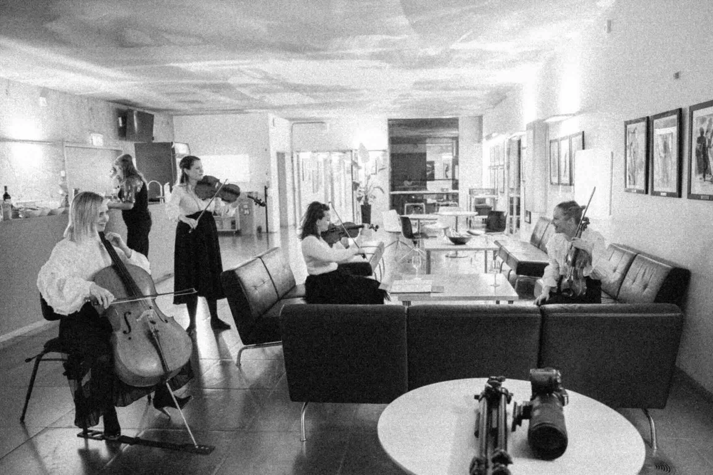 A group of women playing various string instruments, including violins and cellos, in a room decorated with framed artwork on the walls. Some women are standing, while others are seated, as they perform music in a casual setting.