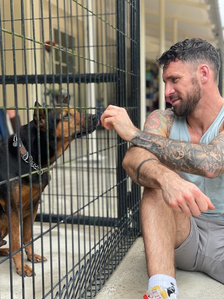 A man with tattoos on his arms squatting next to a dog in a kennel, reaching his hand through the bars to pet or feed the dog, which has a collar and chain leash.
