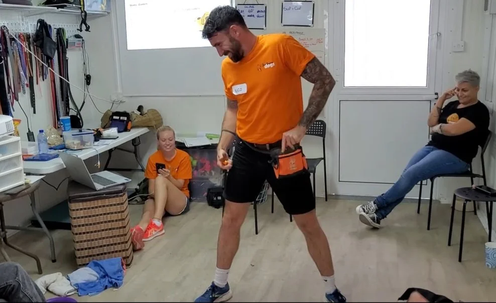 A man in an orange shirt and black shorts laughing while holding a tool in a cluttered room, with two women sitting nearby, one on the floor and the other on a chair, both smiling.