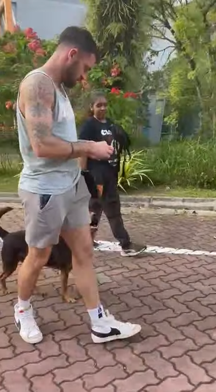 A man with tattoos wearing a sleeveless shirt and shorts walking a dog on a brick pathway outdoors, with a woman and greenery in the background.