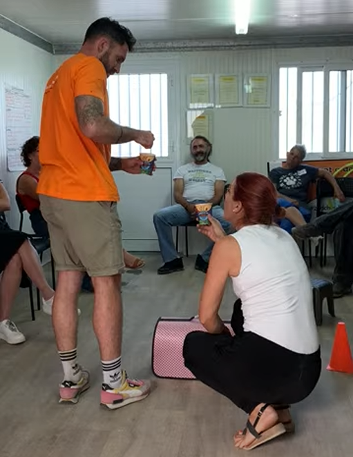 A man in an orange shirt and shorts offers a snack to a woman with red hair kneeling on the floor inside a small room with several seated people. The room has windows and posters on the wall.