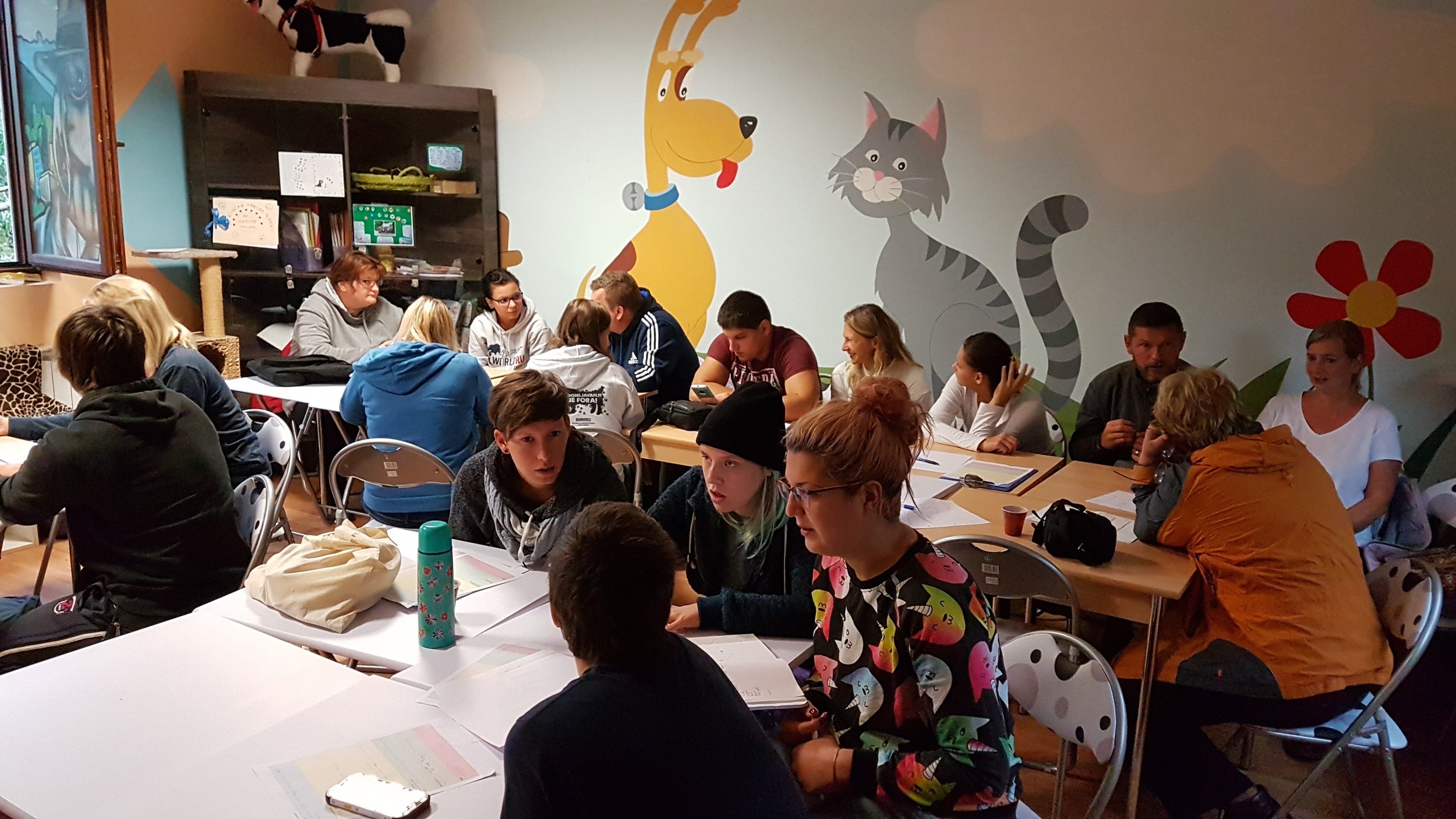 A group of young people gathered around tables in a room with colorful animal wall murals, engaging in discussion or study.