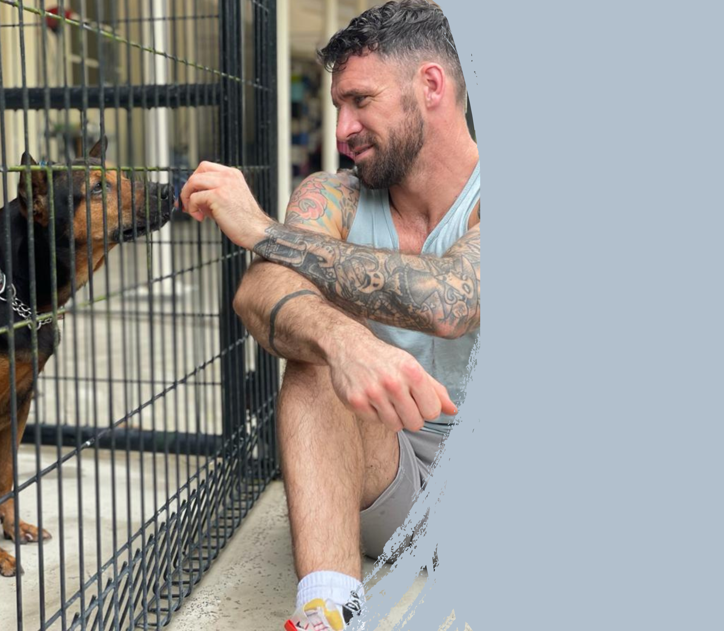 A man with tattoos on his arms is sitting on the ground beside a metal pet cage. Inside the cage, a small brown and black dog is looking at the man while he gently interacts with it.