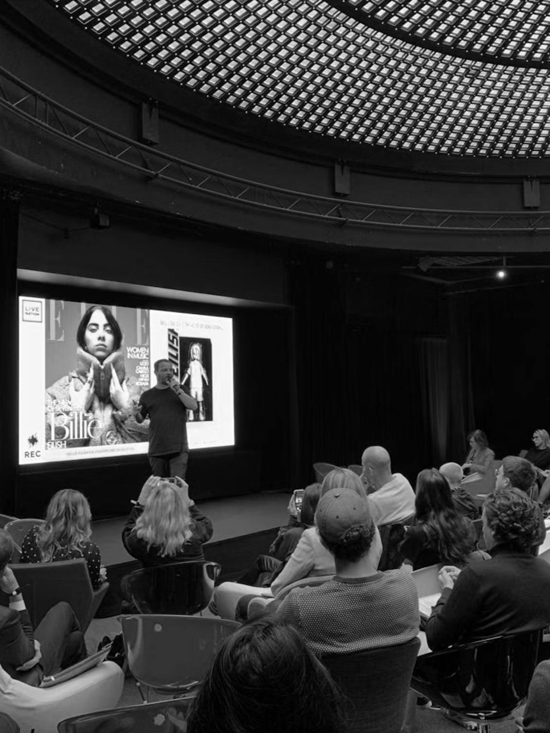 A man giving a presentation on stage in front of an audience, with a large screen behind him displaying an image of Billie Eilish and magazine covers.