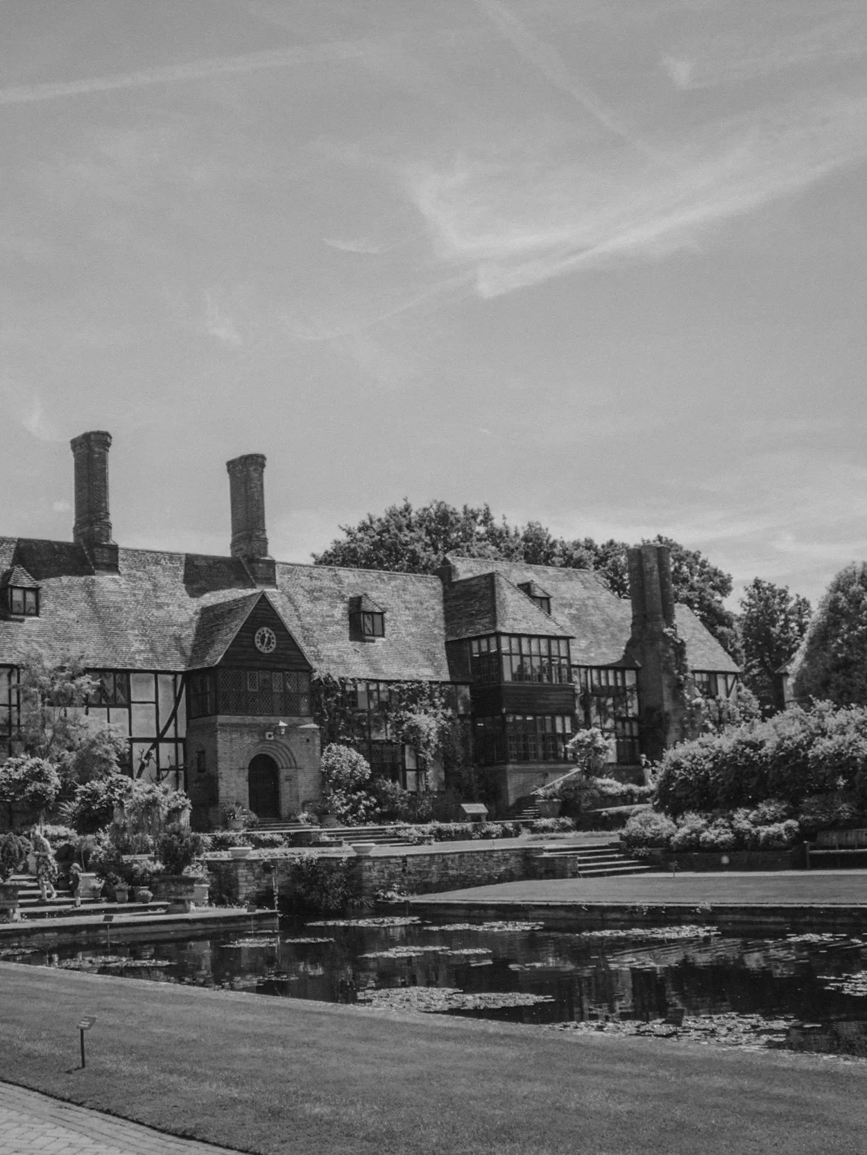 Black and white photograph of a historic mansion surrounded by trees, with a pond and garden in the foreground and a partly cloudy sky above.