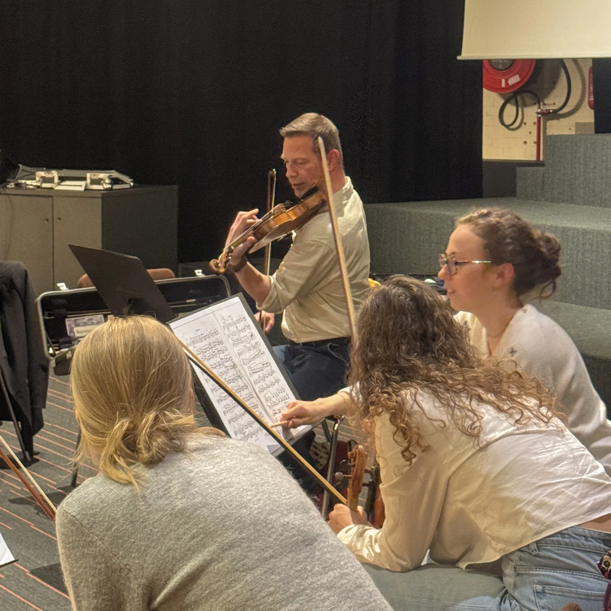 A man playing a violin in front of an audience in a room with musical stands and sheet music, with attendees seated and listening.