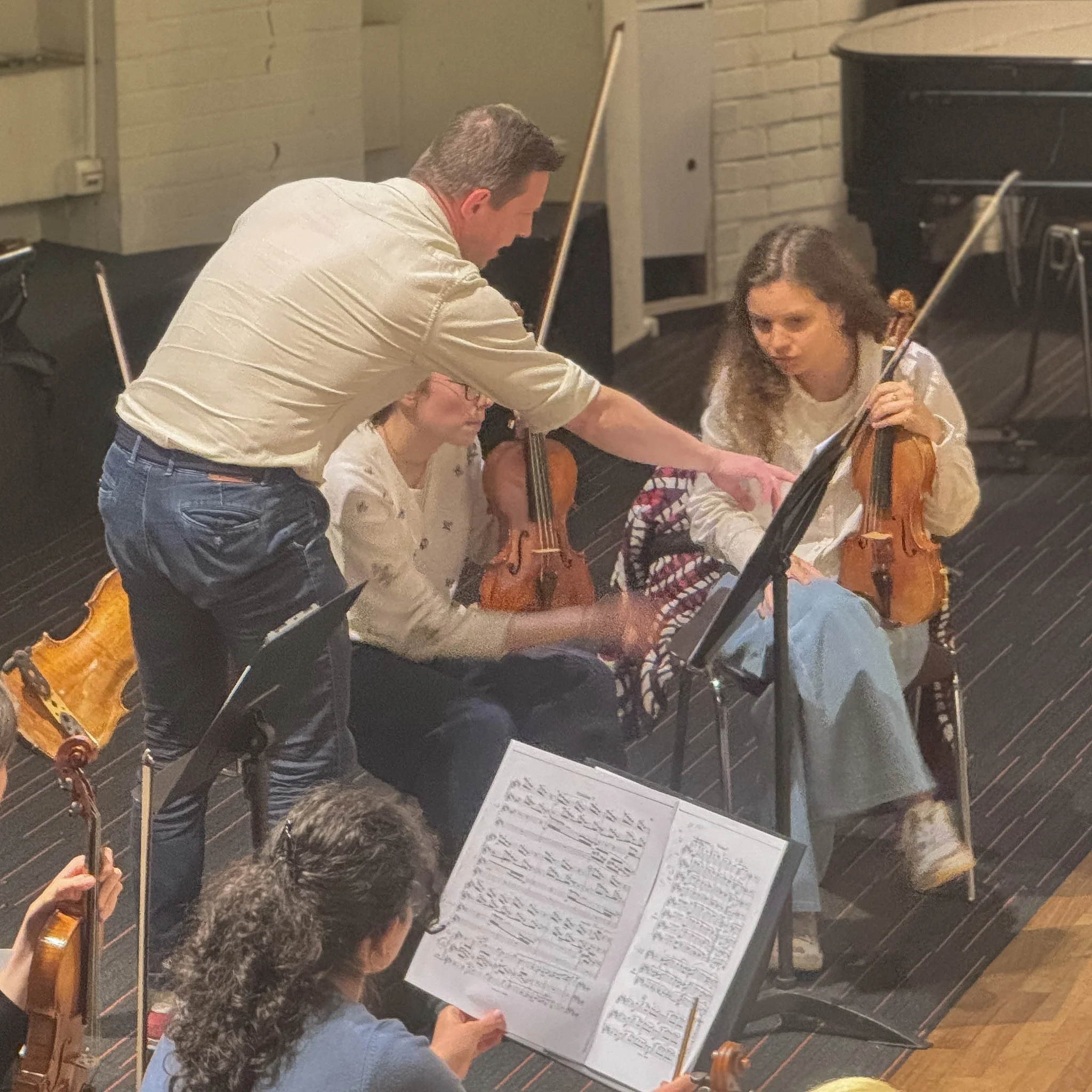 People sitting in chairs, reading music sheets, with a person standing and explaining, possibly in a classroom or rehearsal space with music stands.