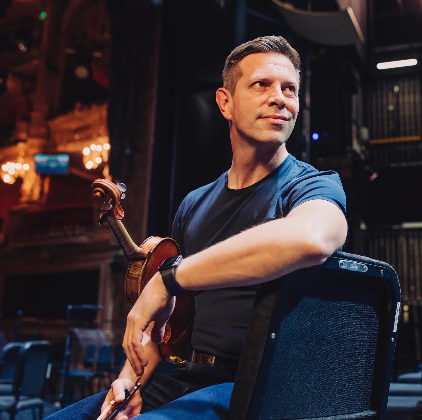 A man sitting on a chair in a dimly lit room holding a violin, looking thoughtfully to the side.