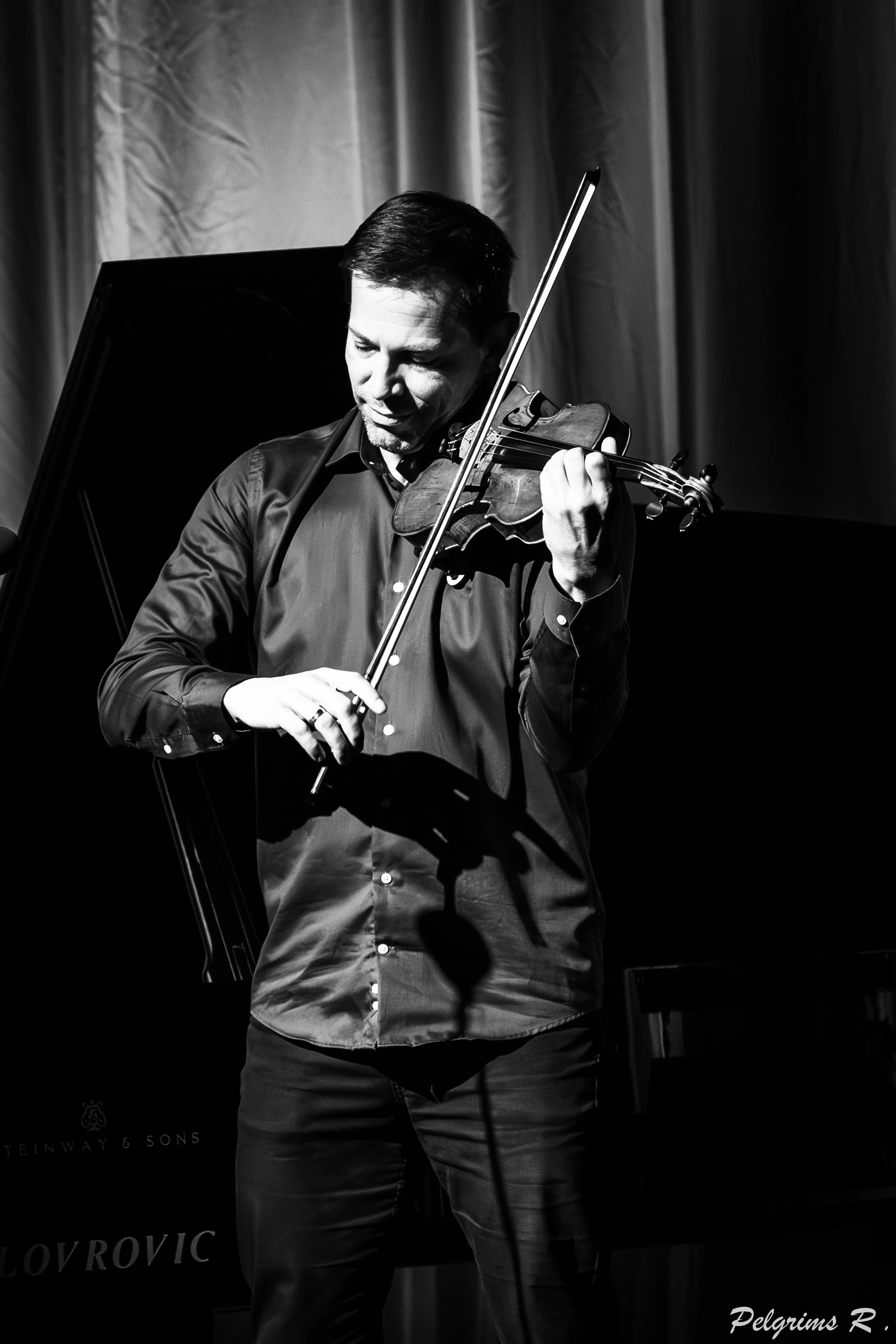 A man playing the violin on stage, dressed in a dark, button-up shirt and pants, with a curtain backdrop.