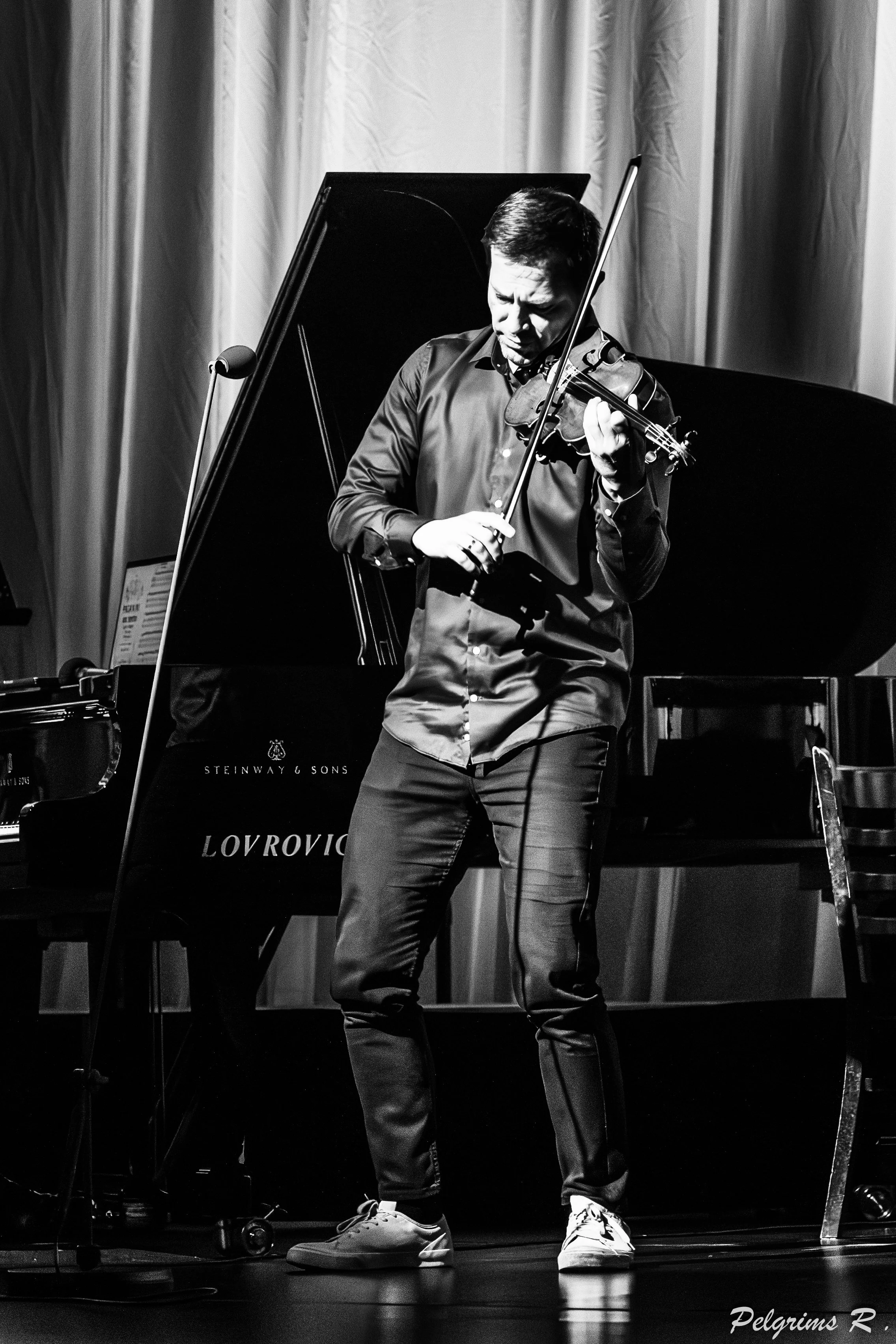 A man playing the violin on stage in front of a piano. The man is wearing a dark shirt, light-colored shoes, and is focused on his performance. The background includes curtains, and the scene is in black and white.