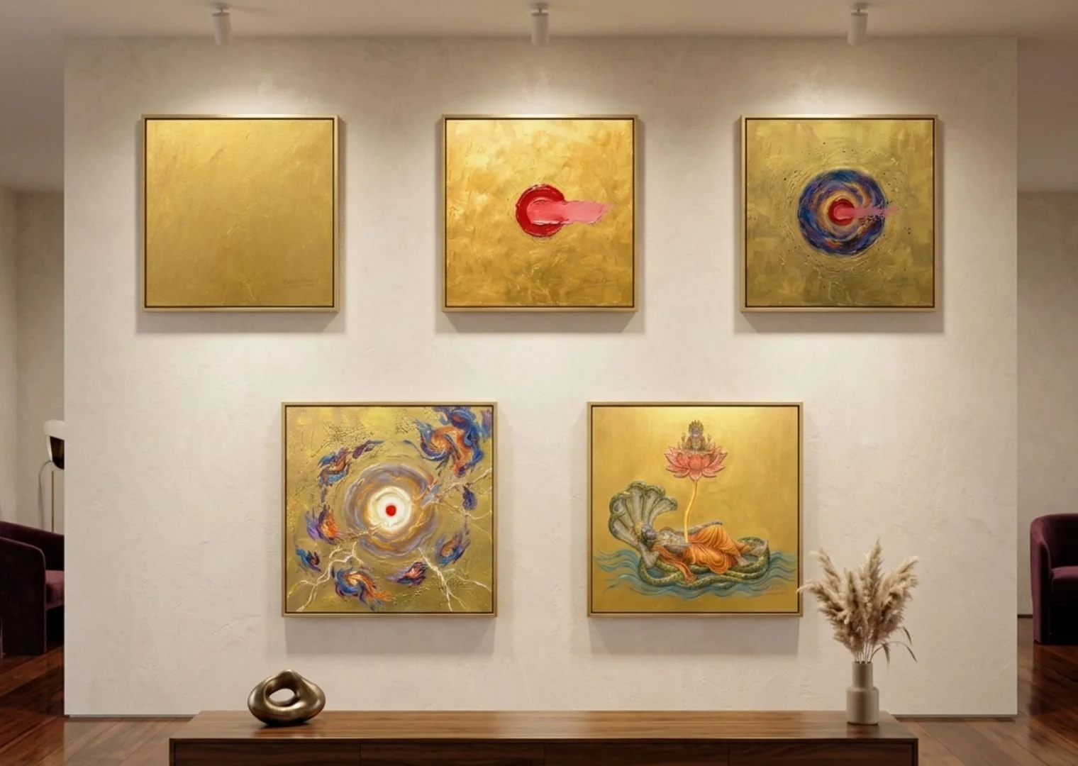 Six abstract paintings with gold backgrounds arranged on a white wall in an art gallery. Some paintings feature colorful circular and cloud-like shapes, with one including a lotus flower and serpent. A wooden console with a decorative sculpture and a vase of dried pampas grass is in the foreground.
