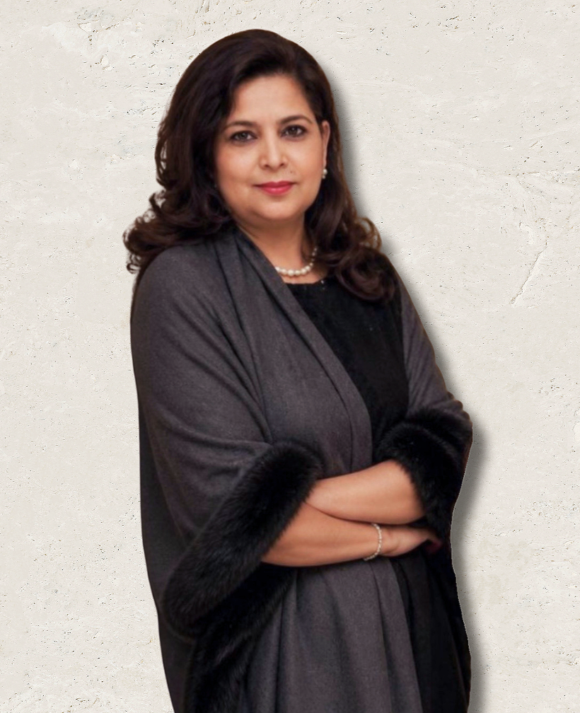A woman with shoulder-length dark brown hair, wearing a pearl necklace, pearl earrings, a gray shawl with black fur trim over a black top, standing with arms crossed against a beige textured background.