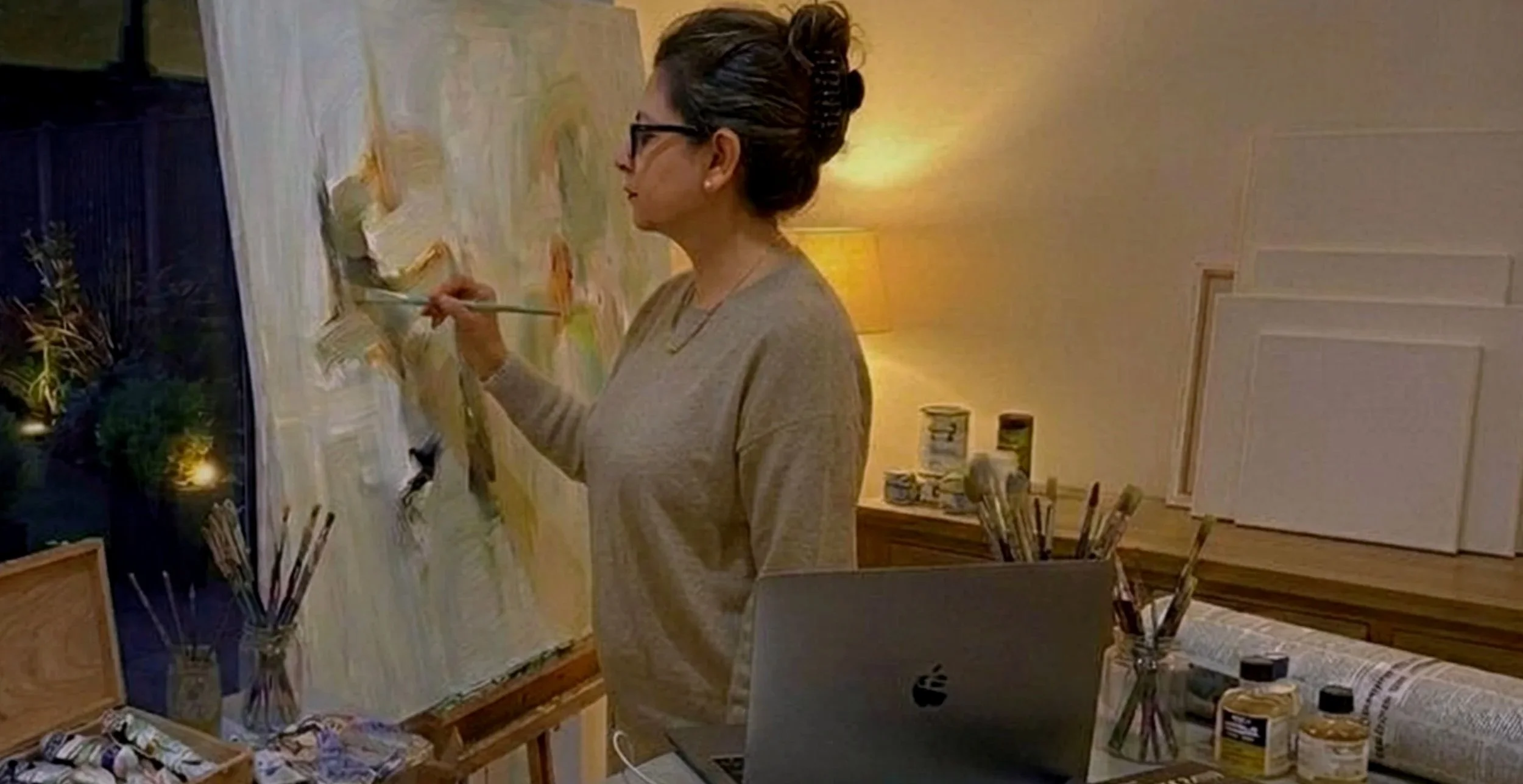 A woman with short hair and glasses is painting a landscape on a large canvas in a cozy room with warm lighting. She is holding a paintbrush and working on a scene featuring a house and trees. A laptop, various paint supplies, and brushes are on the table and surrounding area.