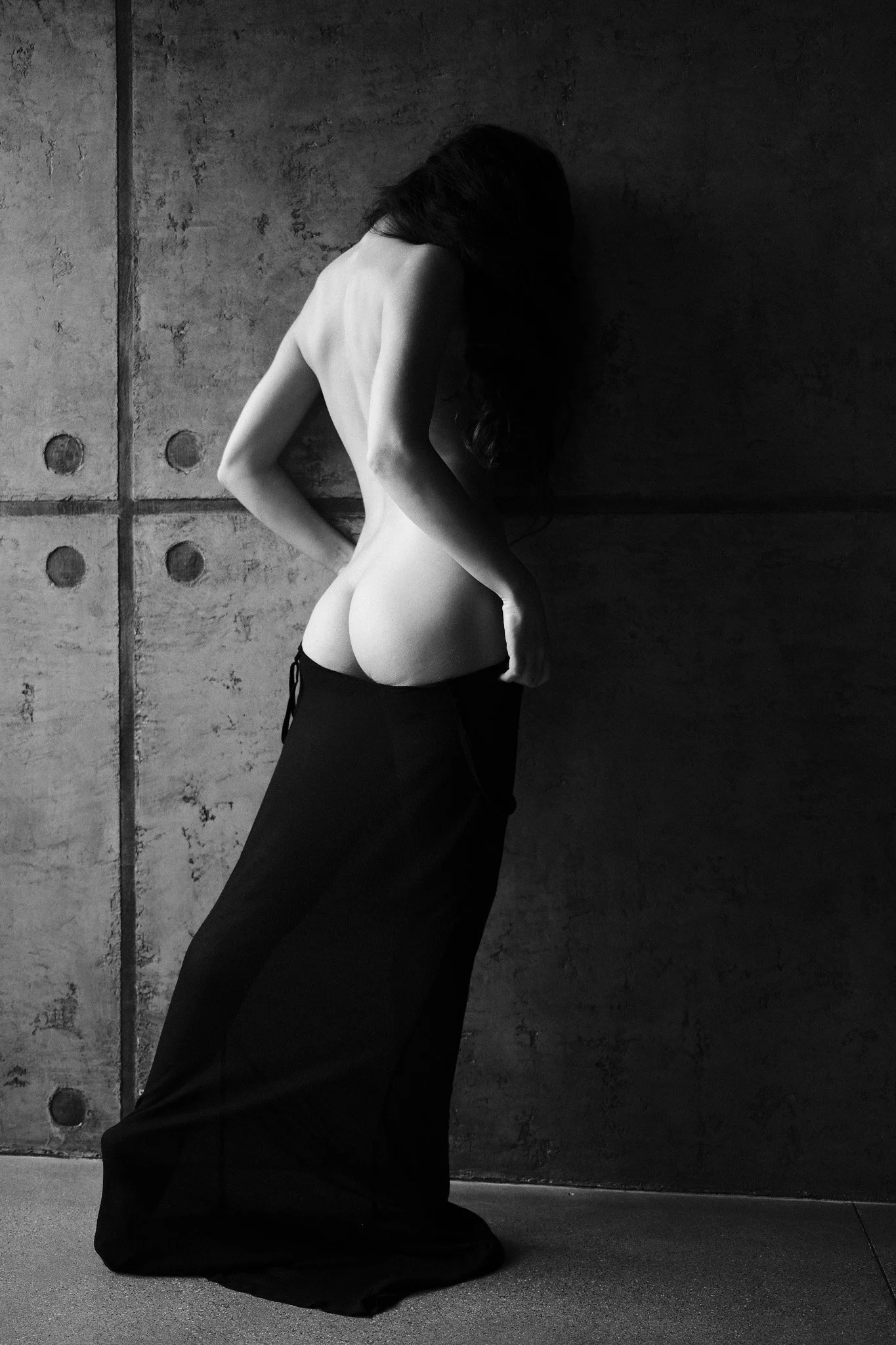 A black and white photograph of a nude woman with long hair, kneeling on the ground against a textured concrete wall. She is facing the wall with her head bowed and her arms resting on her thighs, with her face obscured by her hair.