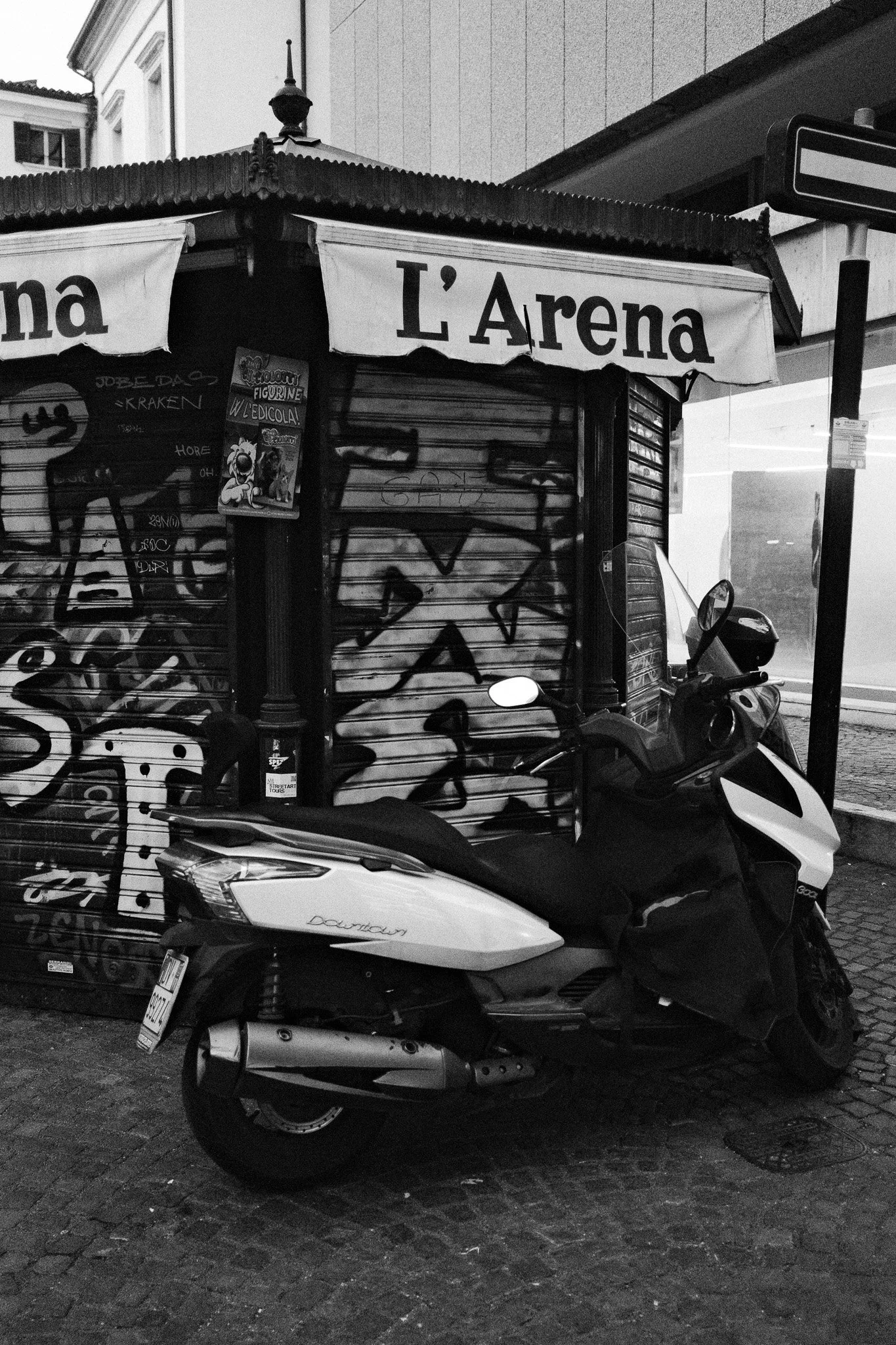 Black and white photo of an outdoor scene featuring a scooter parked on a cobblestone street, with a small booth or kiosk behind it that is graffiti-covered and has a sign reading 'L'Arena' on top. The booth has a corrugated roof and is adjacent to a building.