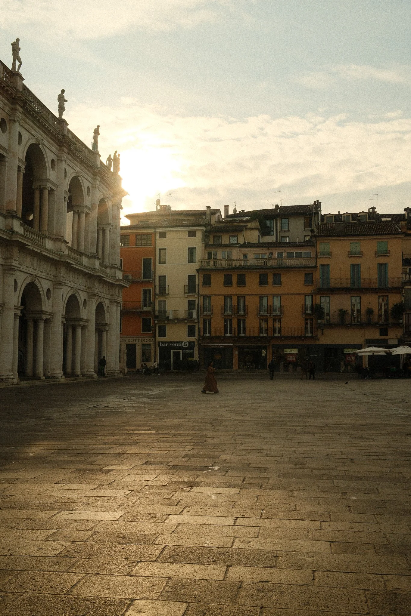 An Italian city square at sunset with historic buildings and a large open cobblestone area, few people walking, and a bright sun partially hidden behind historic architecture.