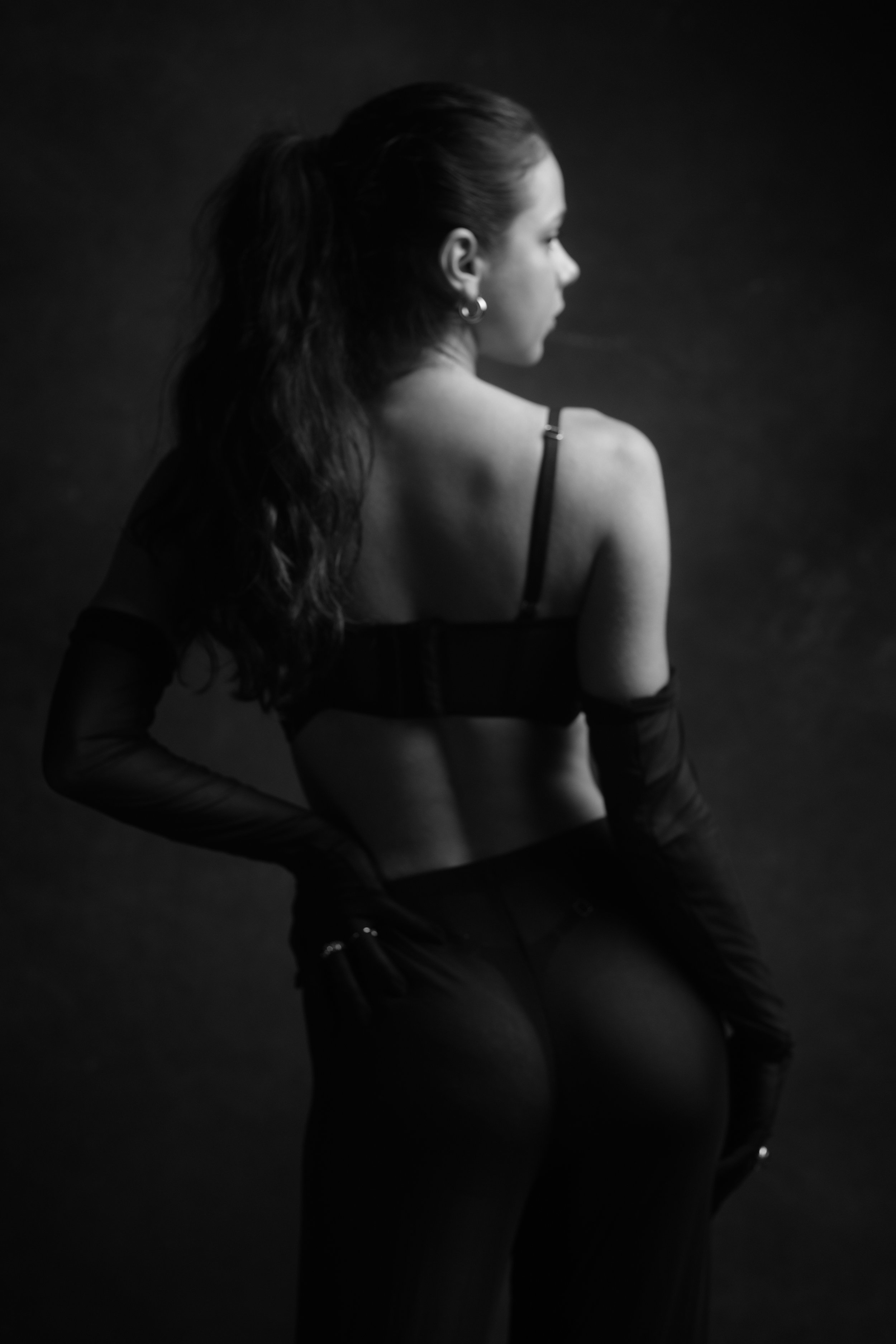 Black and white photo of a woman with long wavy hair wearing a bralette and long gloves, looking away from the camera against a dark background.