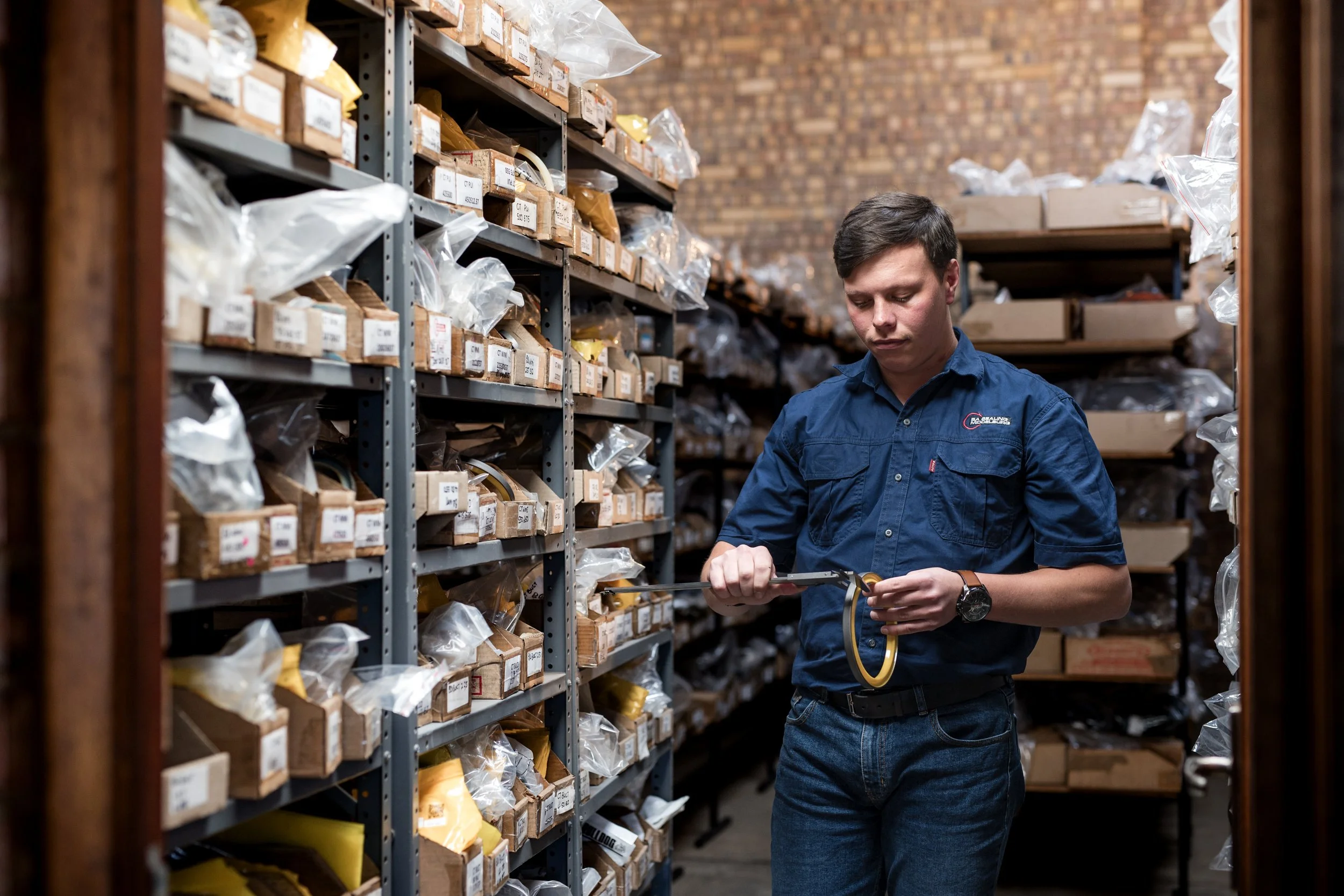 A young man wearing a blue work shirt is inspecting or organizing items on shelves in a storage room or warehouse. The shelves are filled with labeled boxes and plastic bags, and the background shows a brick wall.