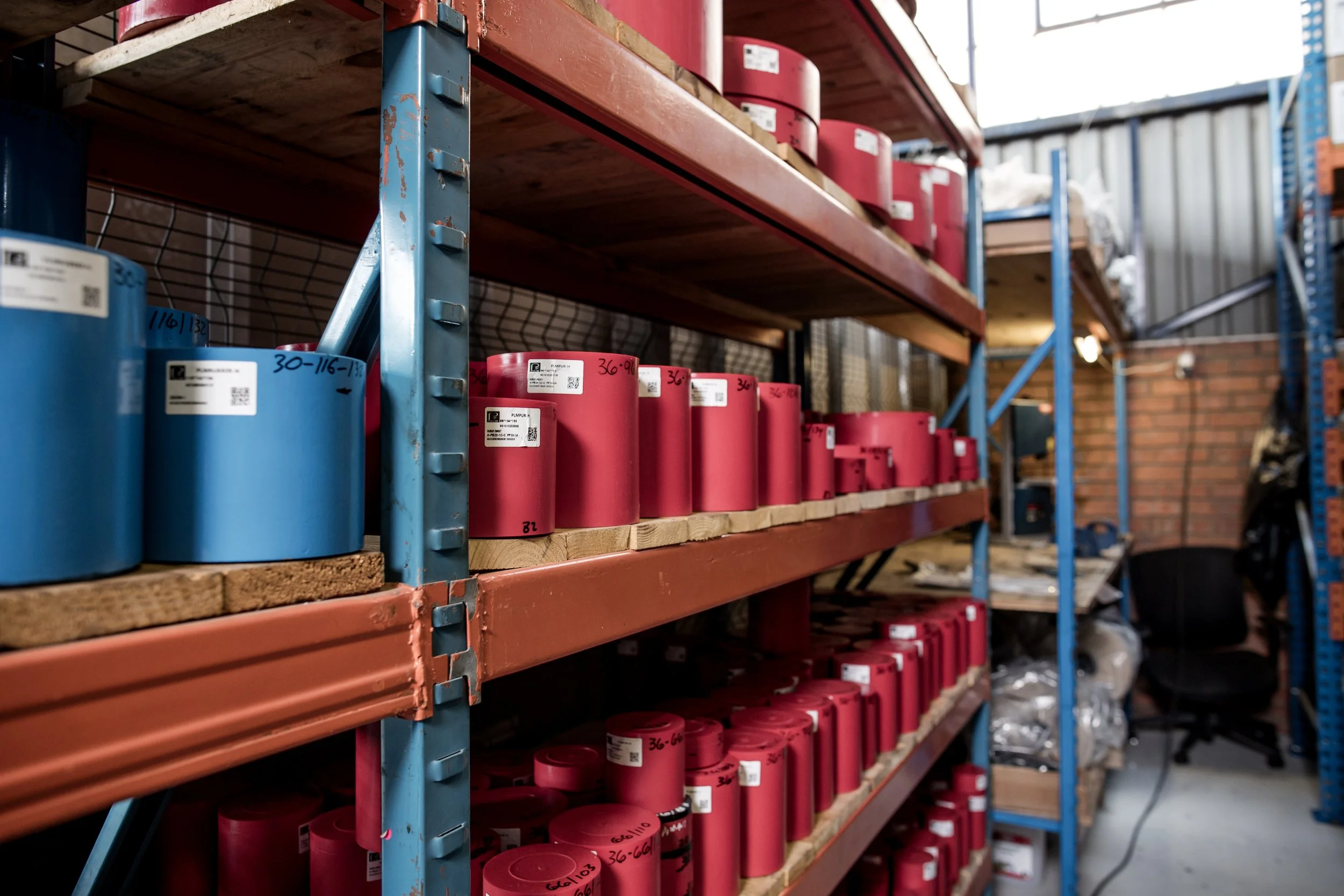 Warehouse shelving with red and blue cylindrical containers labeled with numbers, stored on wooden and metal shelves in an industrial setting.